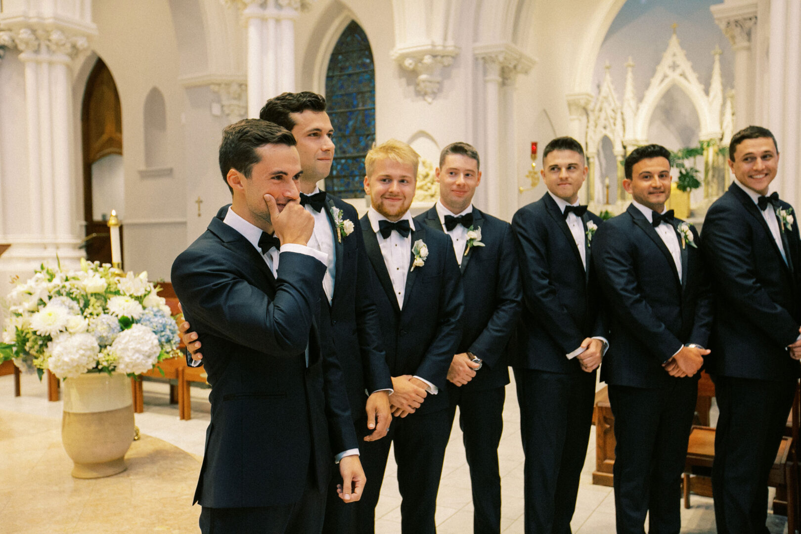 A group of groomsmen in tuxedos stands inside the church at Normandy Farm, smiling and posing for a photo.