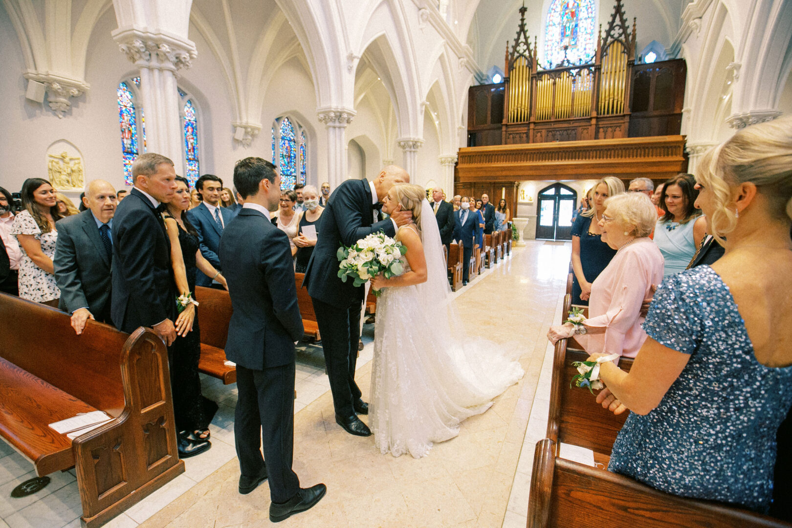 A bride and a man embrace at the altar in a church, surrounded by guests seated in pews. The scene, reminiscent of a Normandy Farm wedding, features stained glass windows and an organ visible in the background.