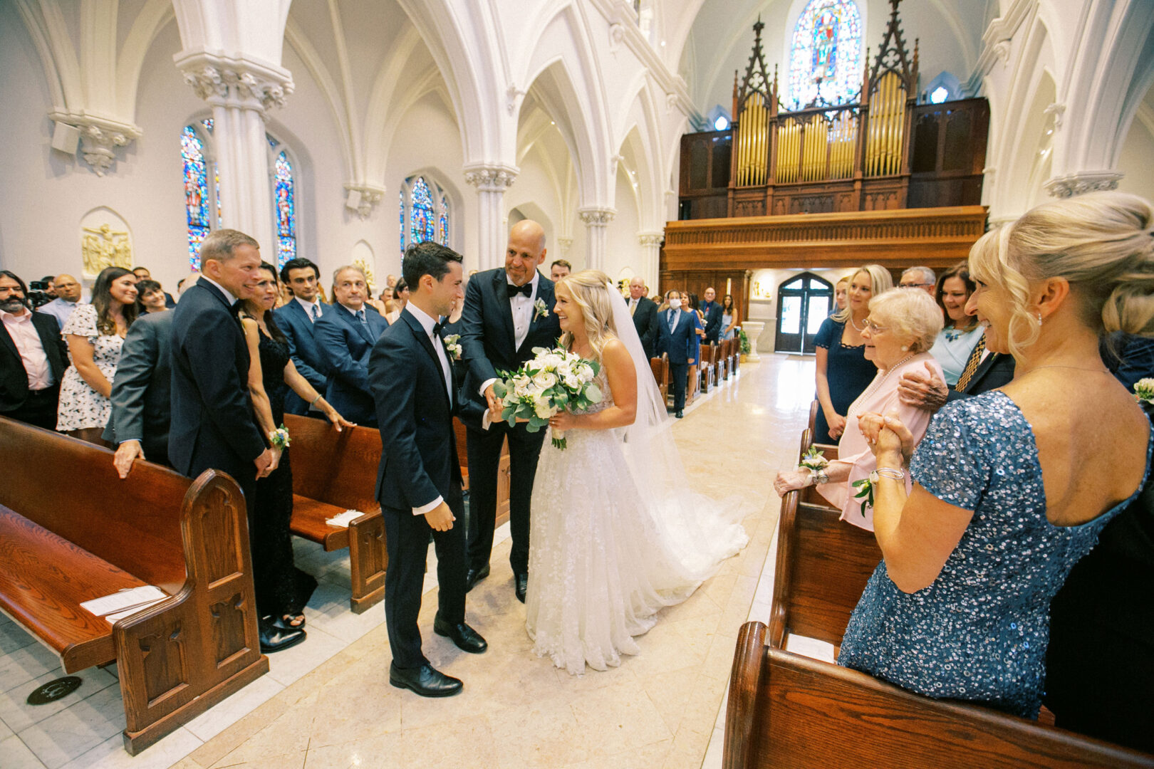 A bride and groom stand at the altar of a charming Normandy Farm wedding, with guests seated in pews. An organ is visible in the background, adding to the rustic elegance.