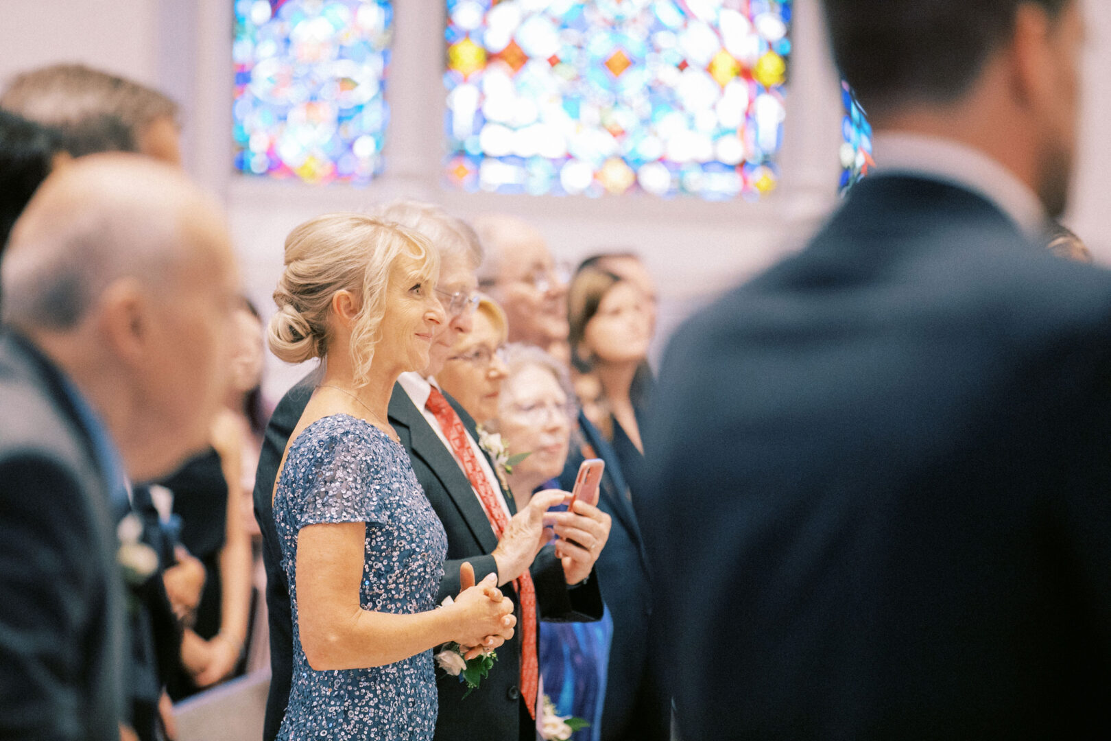 A group of people stands in a church with stained glass windows, dressed in formal attire, reminiscent of a Normandy Farm wedding. One woman holds a phone.