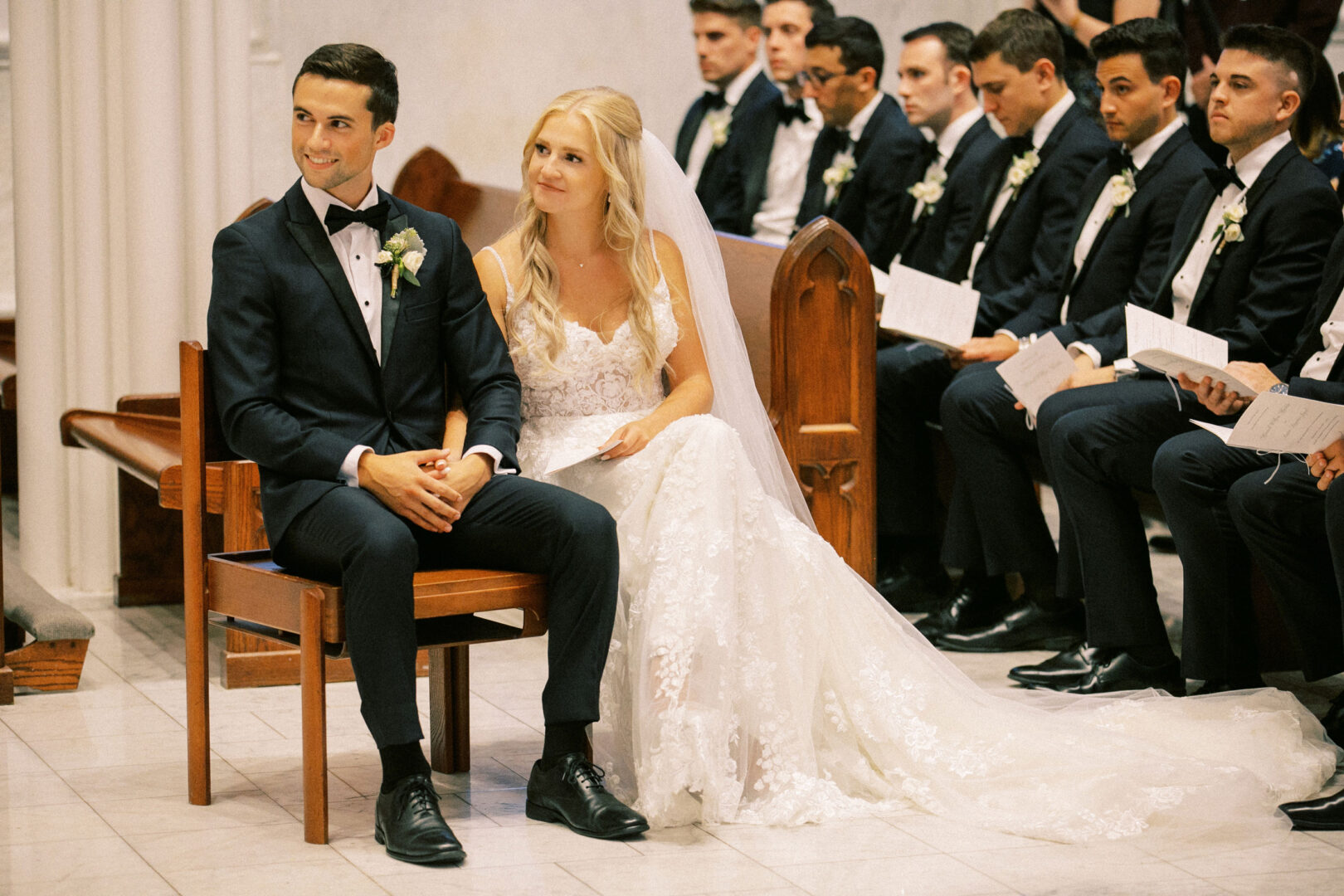 At a picturesque Normandy Farm wedding, a bride and groom sit together during the ceremony, surrounded by groomsmen in dark suits holding programs.