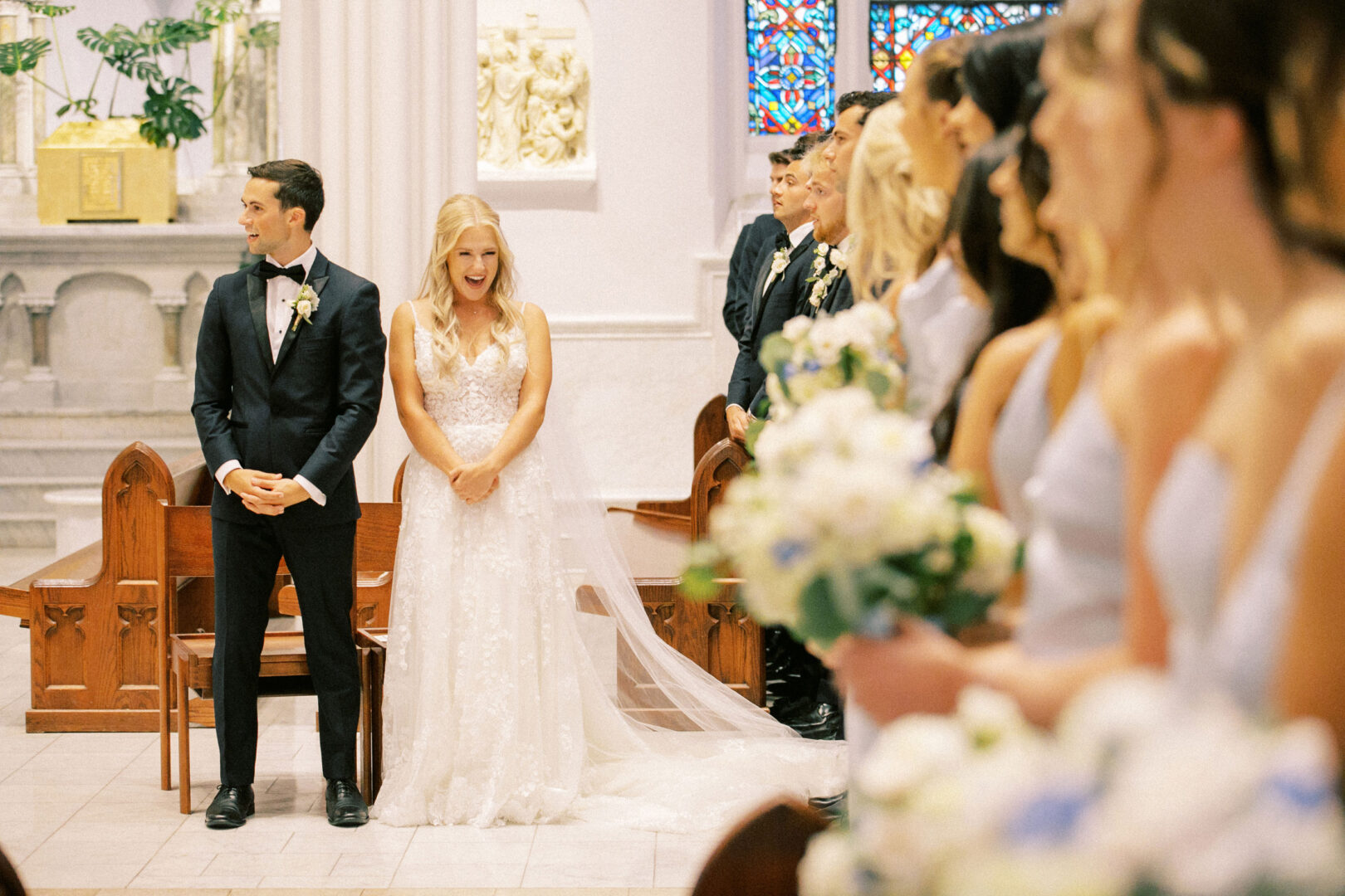 At a charming Normandy Farm wedding, a bride and groom stand together at the altar in a church, with bridesmaids holding bouquets in the foreground.