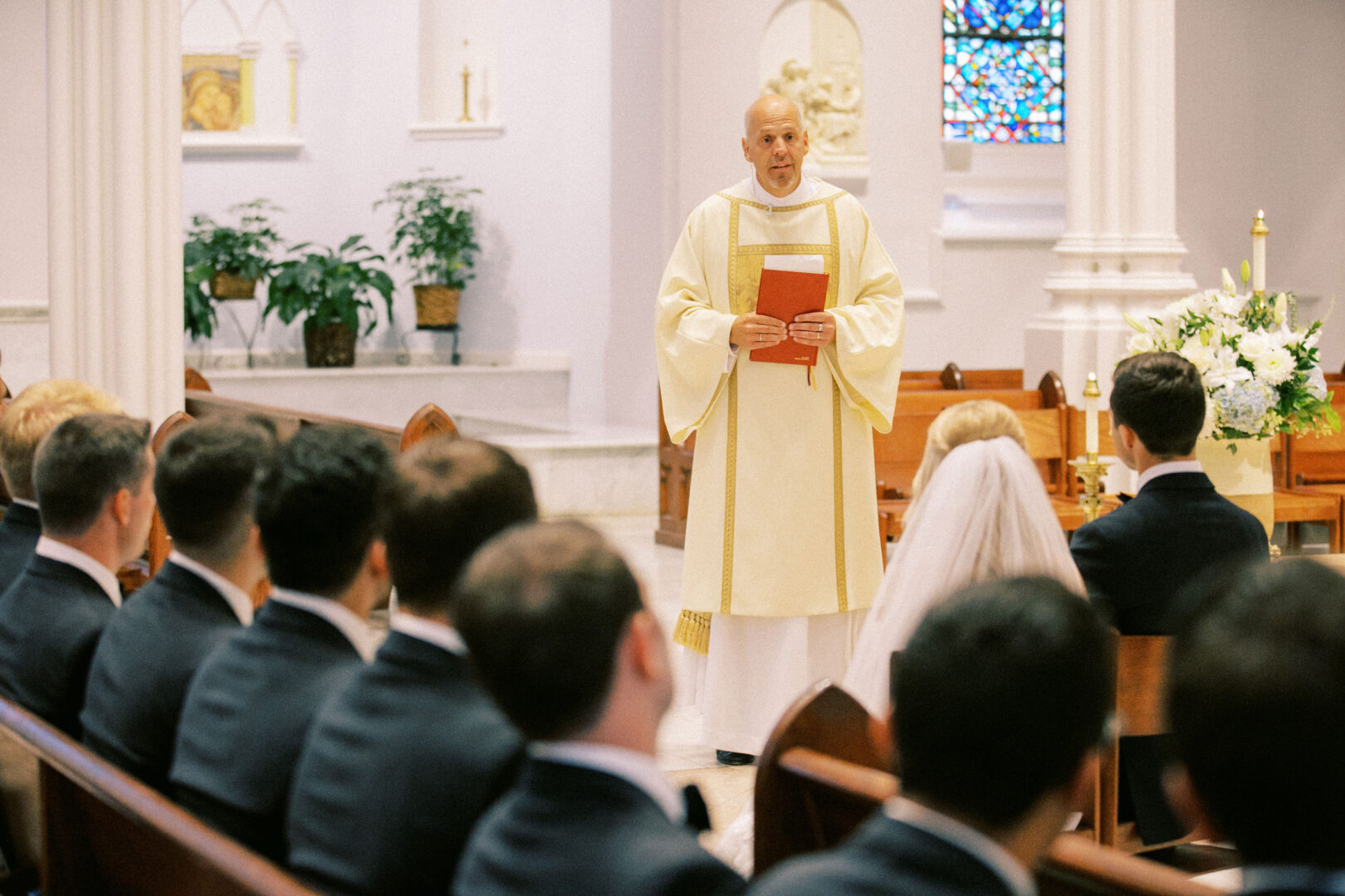 A priest stands at the front of the church at Normandy Farm Wedding, holding a book, with a bride and groom seated before him and several guests watching from pews.