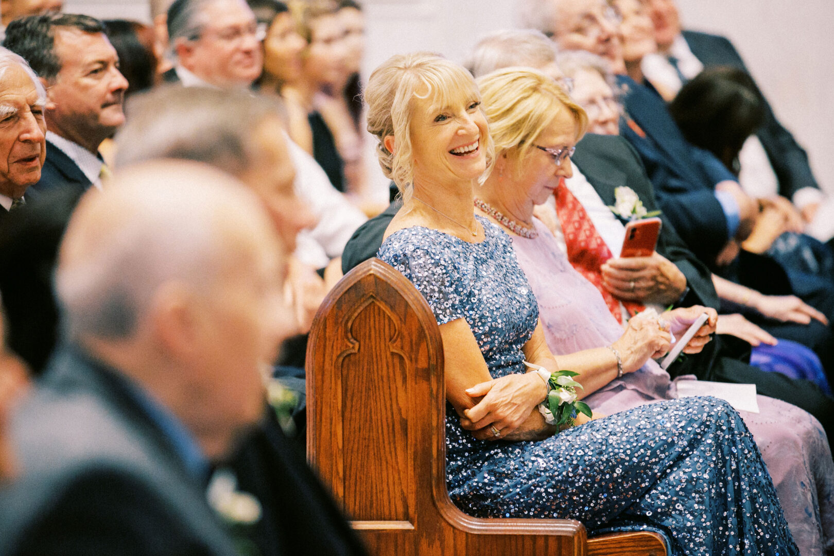 At a Normandy Farm wedding, audience members are seated, with a woman in a blue dress smiling warmly while others are captivated by their picturesque surroundings.