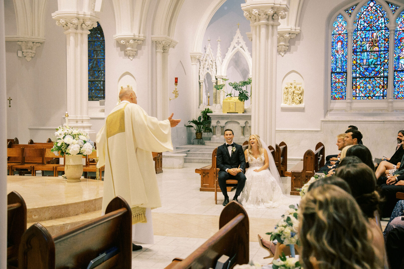 A couple sits during a Normandy Farm wedding ceremony in a church. A priest in white robes stands before them. Guests are seated nearby, and stained glass windows decorate the background.