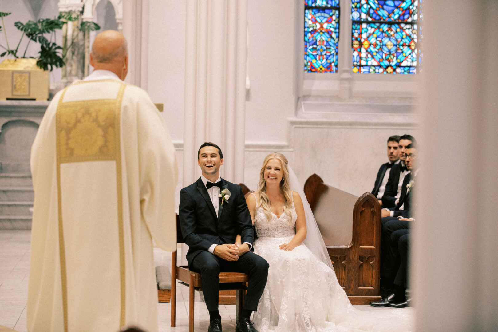 A bride and groom sit smiling during their Normandy Farm wedding ceremony in a church, with a priest standing nearby and stained glass windows in the background.