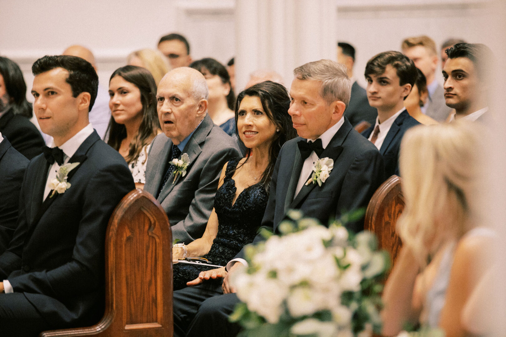 Guests elegantly seated in a church for a Normandy Farm wedding; the atmosphere is filled with sophistication as attendees don suits and dresses, surrounded by exquisite floral arrangements that enhance the charm of this formal event.