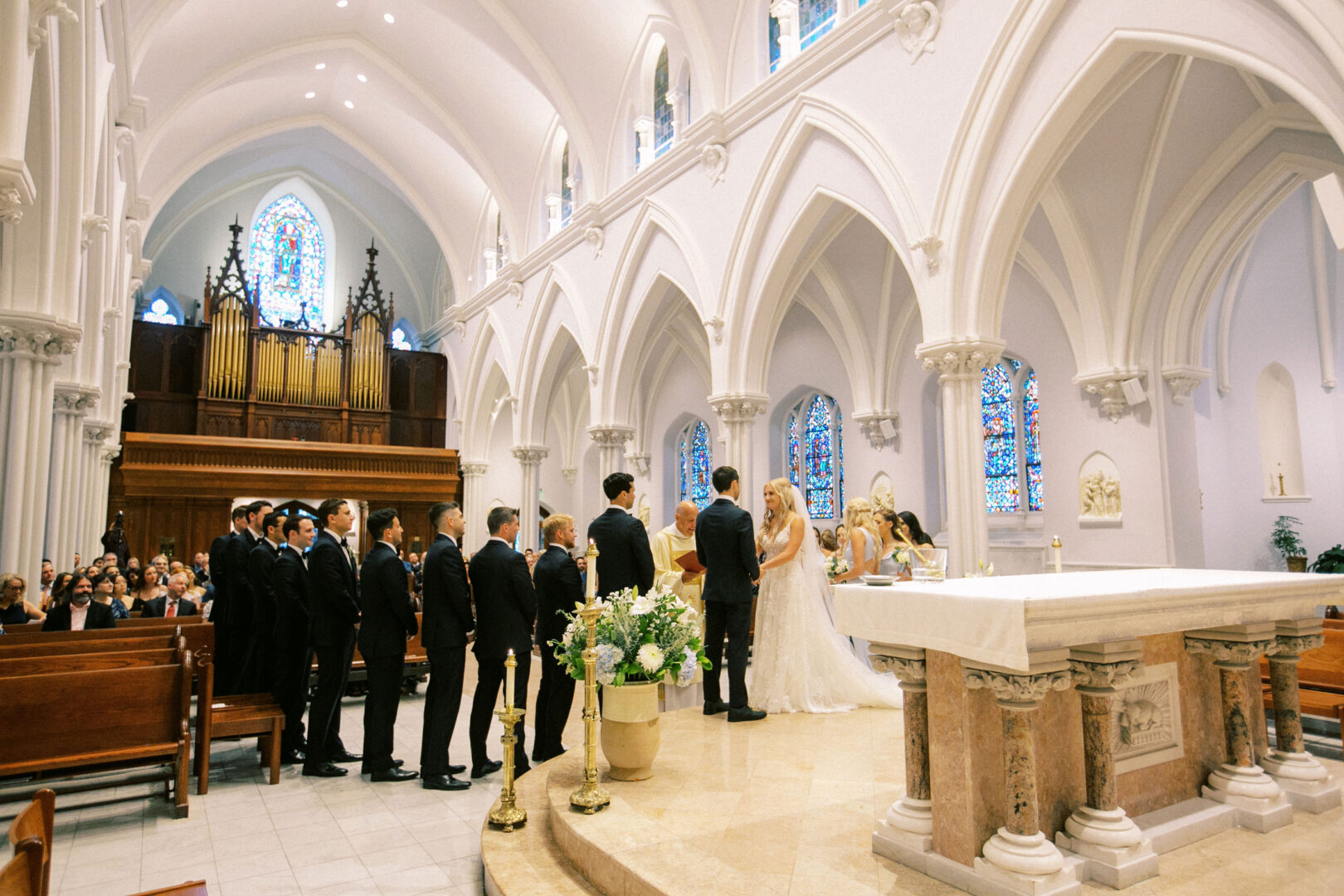 A couple stands at the altar during a Normandy Farm wedding ceremony, framed by church's stained glass windows. The bridal party gathers close, their joy mirrored by guests seated in pews.