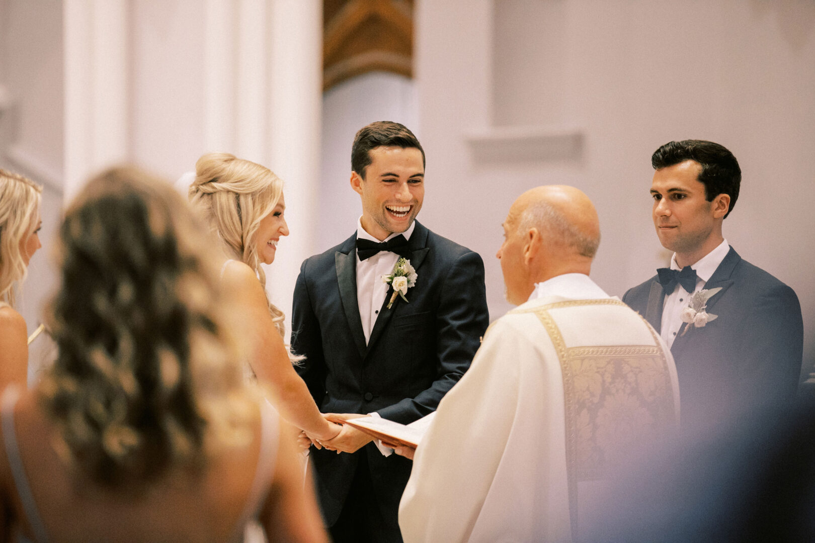 A couple exchanges vows at Normandy Farm, smiling at each other while a priest officiates and guests look on, creating a picturesque wedding scene that captures hearts.