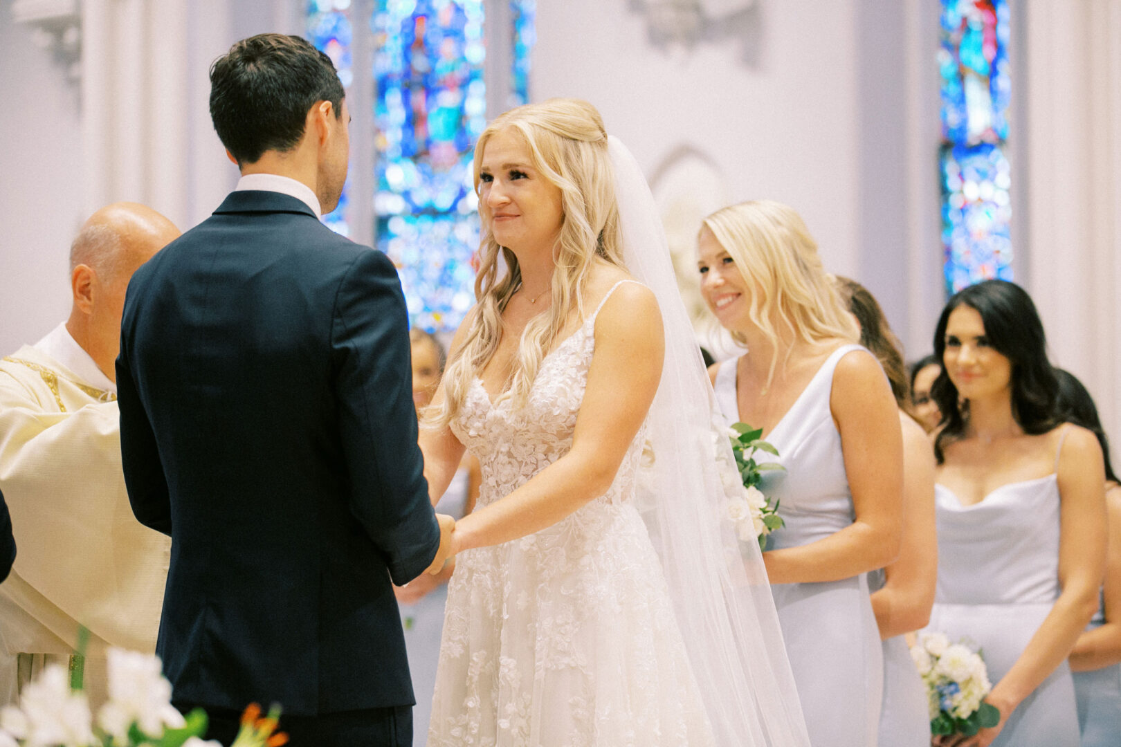 A bride and groom stand facing each other during a picturesque Normandy Farm wedding ceremony in a church with stunning stained glass windows, accompanied by bridesmaids in light dresses.