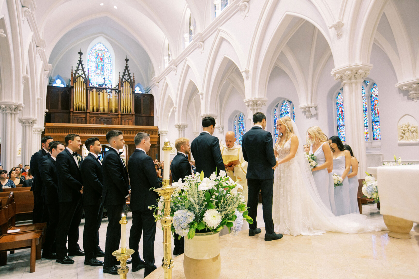 A Normandy Farm wedding unfolds beautifully as the bride and groom stand at the altar, surrounded by bridesmaids and groomsmen. The serene church interior, adorned with stunning stained glass windows and an ornate organ, adds to the charm of this unforgettable day.