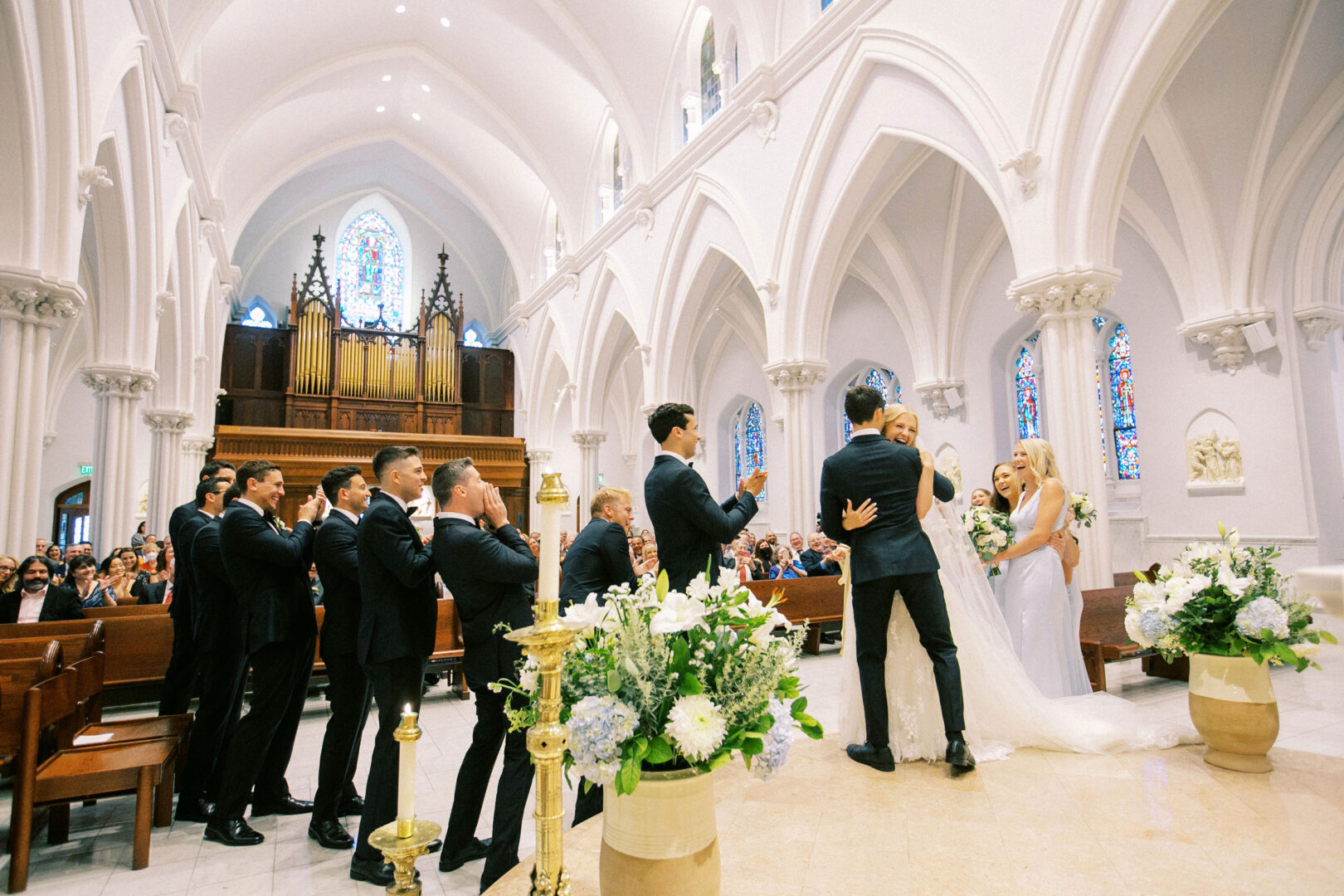 A couple shares a kiss at the altar in a charming church, reminiscent of a Normandy Farm Wedding. Groomsmen and bridesmaids clap nearby, surrounded by the glow of stained glass windows and the gentle sound of an organ.