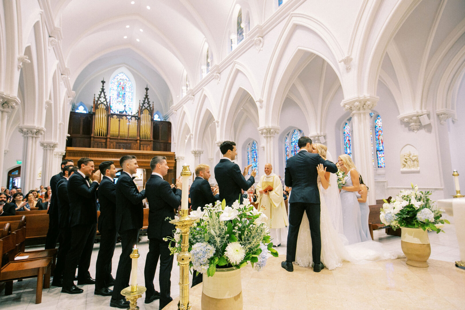 A couple exchanges vows in a church with an ornate interior; groomsmen and a priest stand nearby, and guests are seated, capturing the elegance of a Normandy Farm wedding.