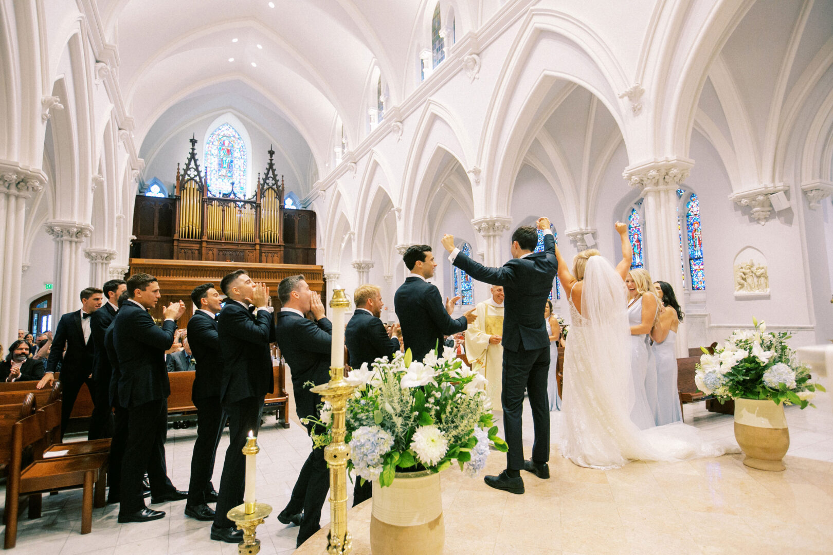 A beautiful Normandy Farm wedding unfolds within a church adorned with stained glass, where the couple stands at the altar. Surrounded by their elegantly dressed wedding party and a priest, the scene is both timeless and deeply romantic.