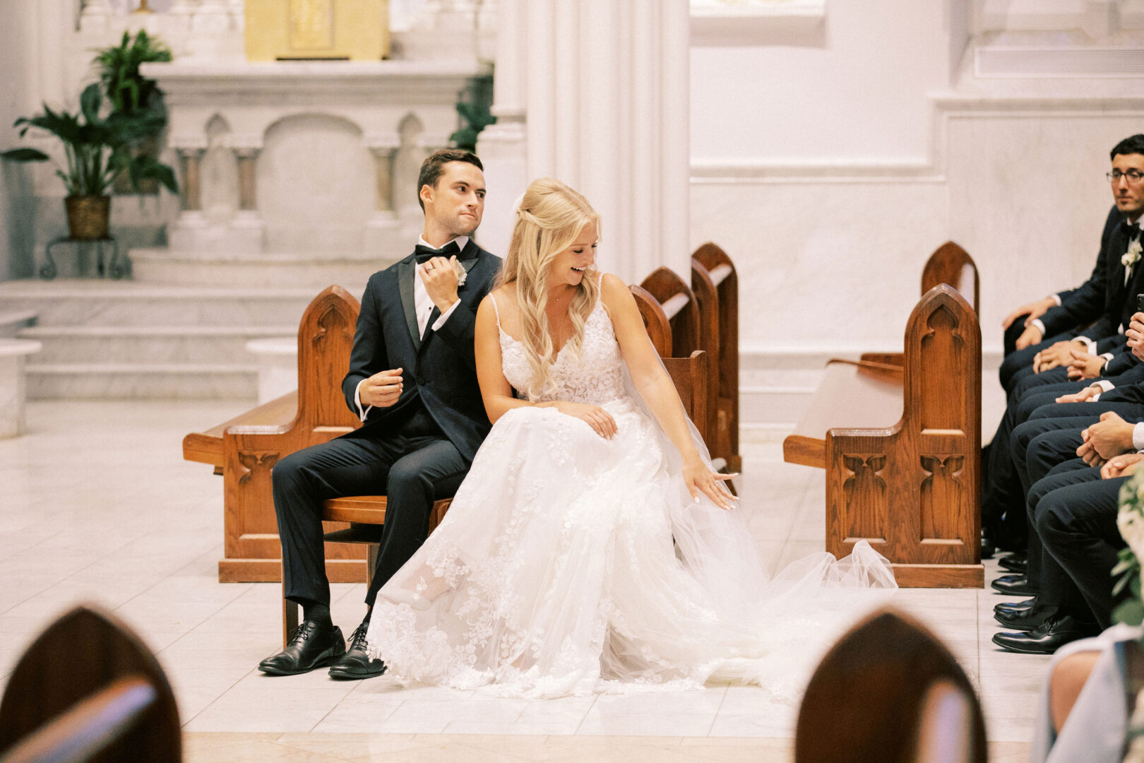 At their Normandy Farm Wedding, the bride and groom sit together at the front of a charming church. She gently straightens her dress as he adjusts his bow tie, while guests are visible seated in the pews, eagerly awaiting their vows.