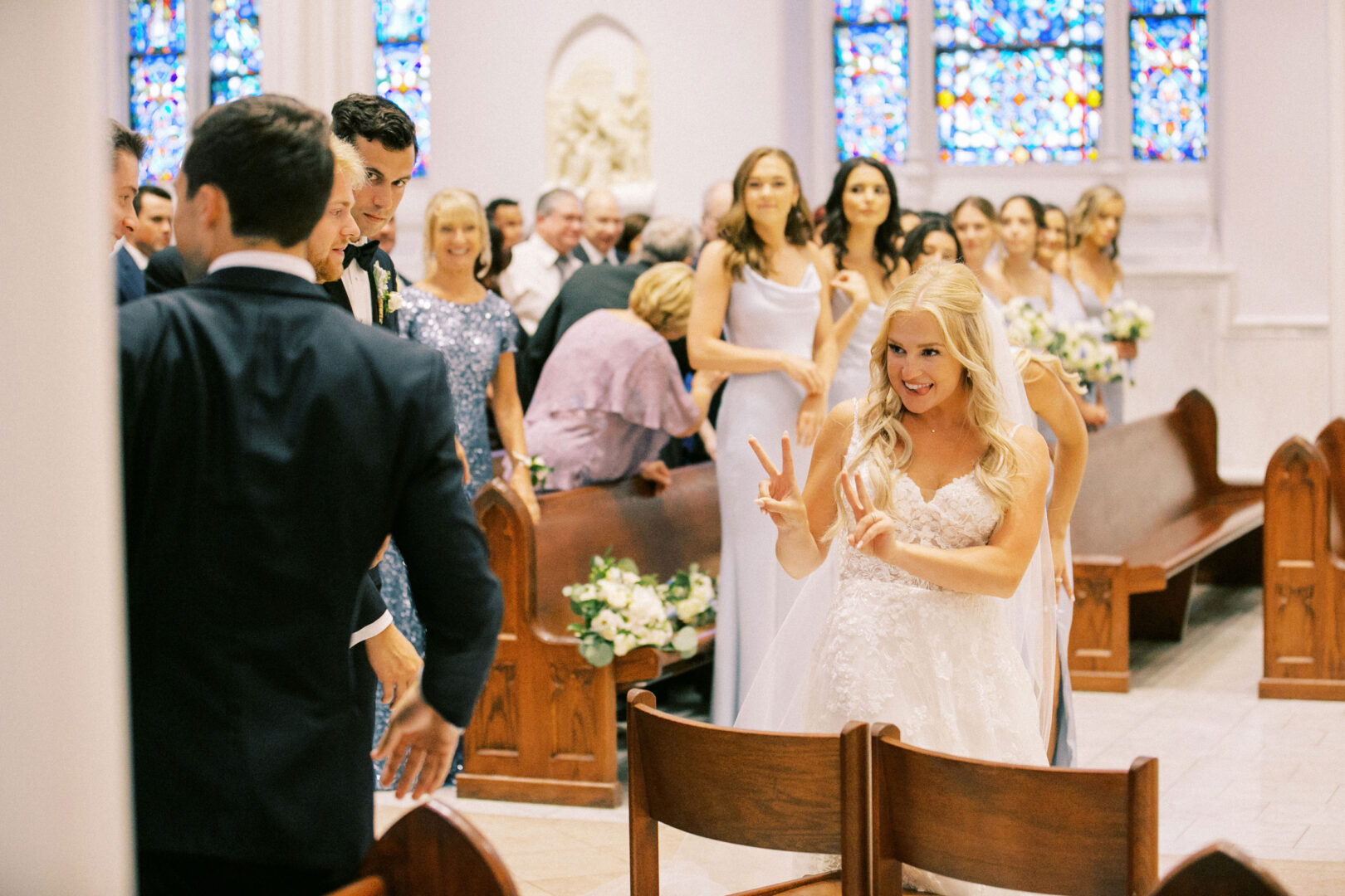 At a charming Normandy Farm wedding, the bride in a white dress playfully gestures with two fingers at a person in a suit during the church ceremony. Guests in formal attire are seated nearby, while stained glass windows beautifully adorn the background.