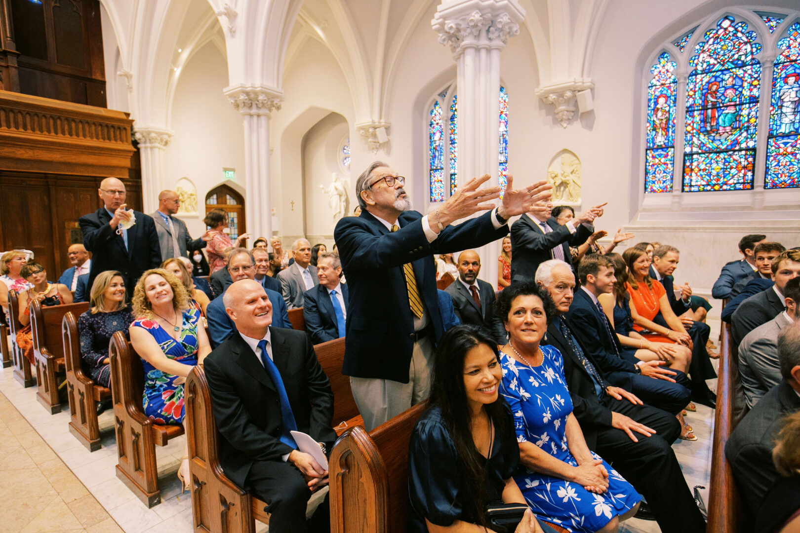 People seated in a church, some standing and clapping, as if celebrating a Normandy Farm wedding. The interior's charm is heightened by the stained glass windows and columns.