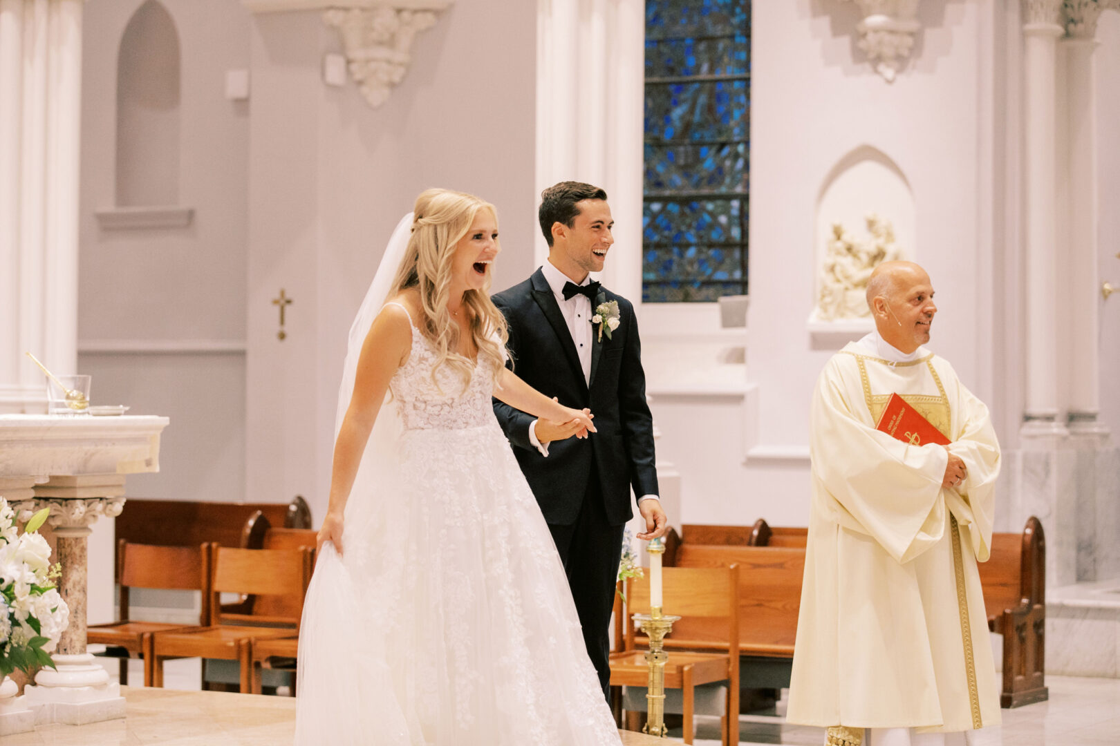 A bride and groom hold hands and smile during their Normandy Farm wedding ceremony in a church, with a priest standing nearby.