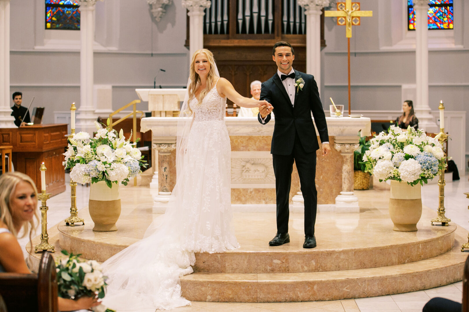 At their Normandy Farm wedding, the bride and groom smile while holding hands at the altar of a church, surrounded by elegant white and blue floral arrangements.