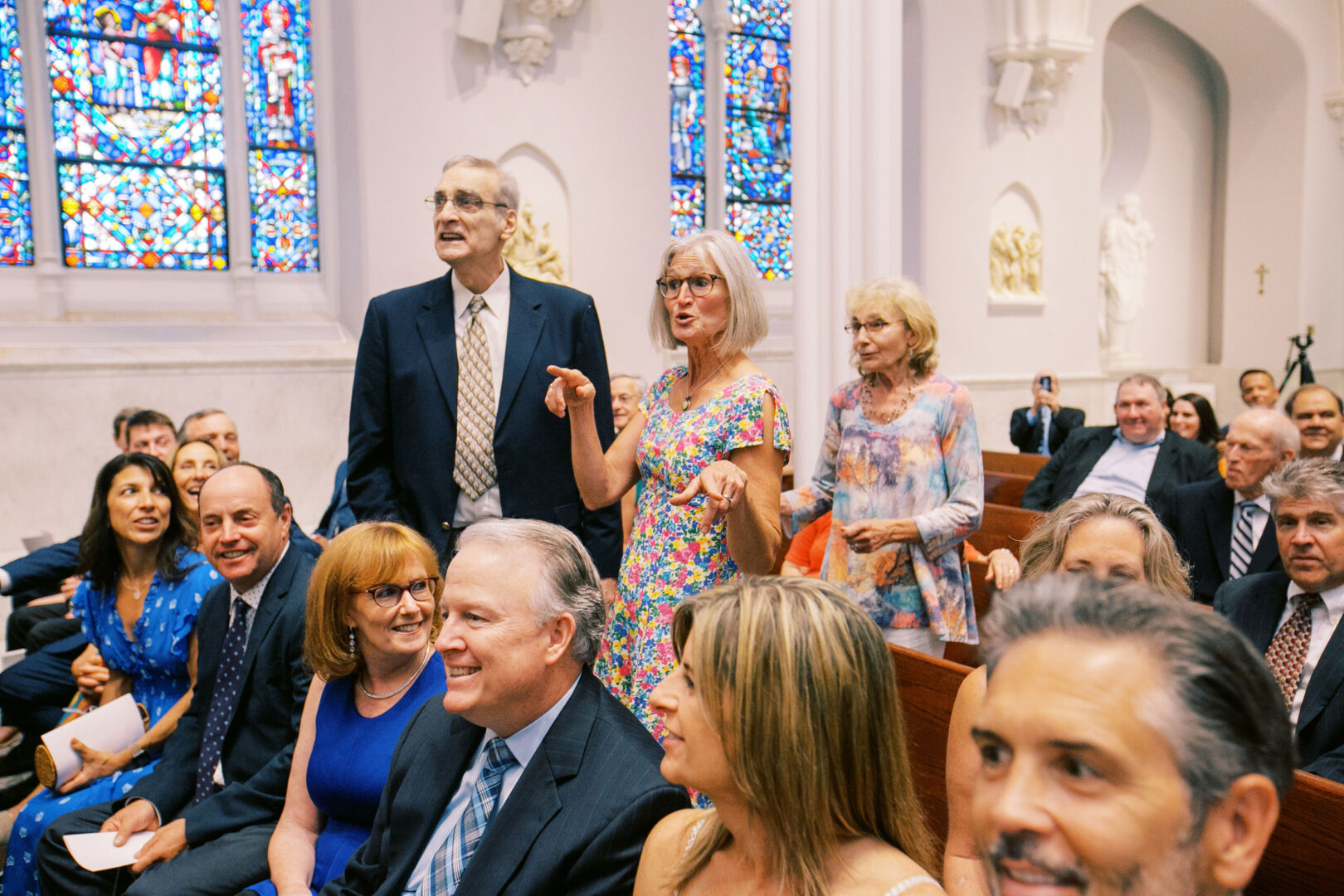 People dressed formally, seated in a church with stained glass windows reminiscent of a Normandy Farm wedding. Some stand and appear to be talking or engaging with others.