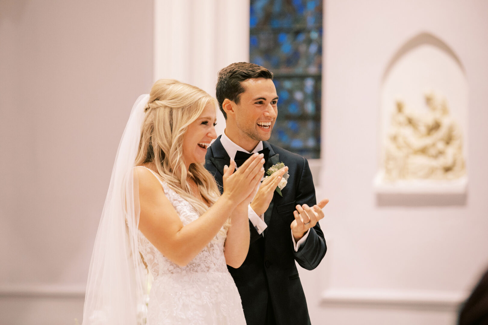 A bride and groom smile and clap during their Normandy Farm Wedding ceremony inside a charming church.