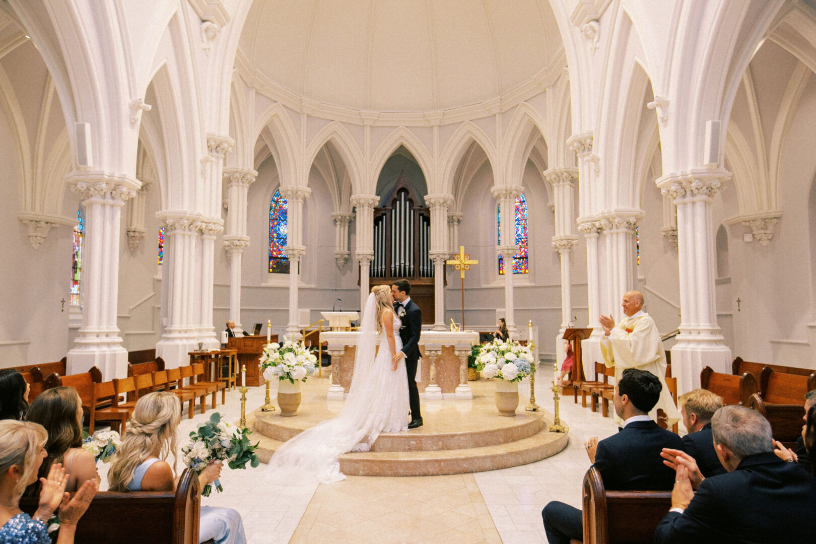 A couple kisses at the altar in a grand church with white arches and stained glass windows, reminiscent of a Normandy Farm wedding. Guests are seated, and a priest stands nearby.