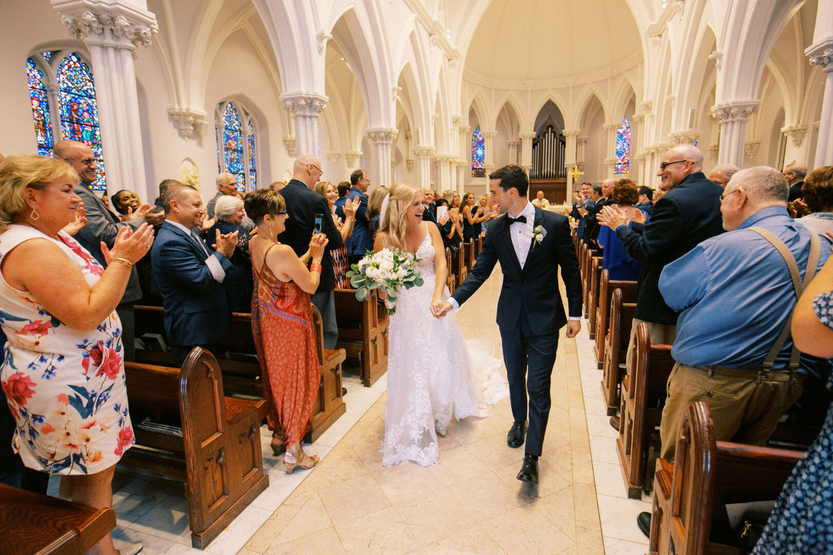 At a charming Normandy Farm wedding, the bride and groom hold hands as they walk down the aisle in a quaint church, surrounded by guests who clap and smile warmly.