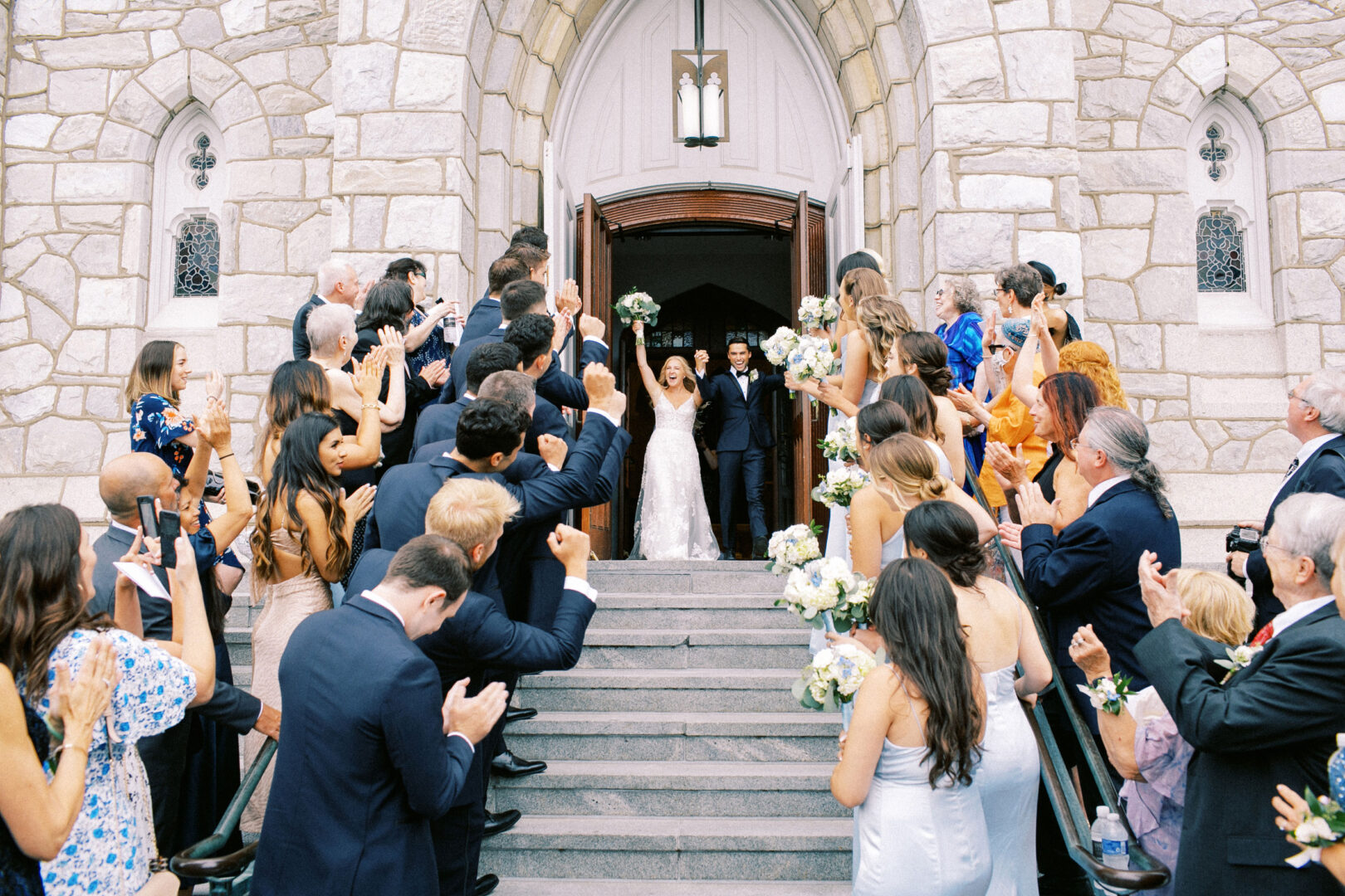 Newlyweds exit a stone church at Normandy Farm, greeted by a crowd of cheering guests holding white flowers.