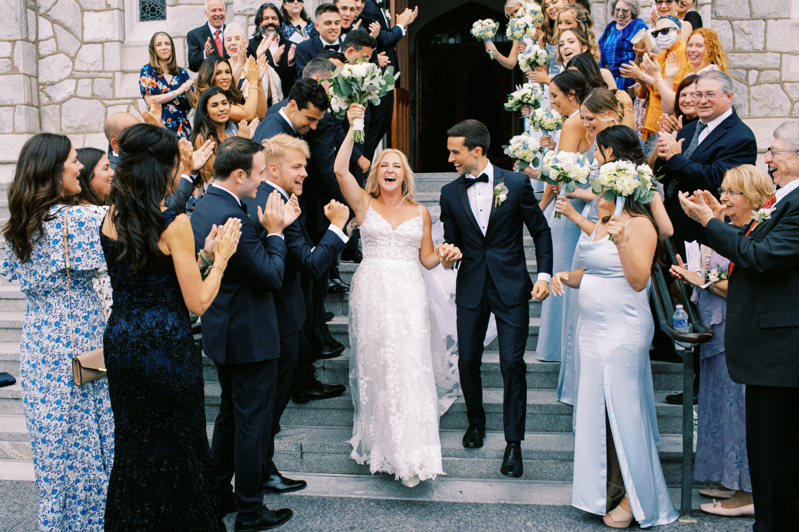 At a picturesque Normandy Farm wedding, a bride and groom celebrate with a cheering crowd on stone steps. She holds a bouquet, and they both smile. Bridesmaids in light blue dresses stand nearby, adding to the joyous atmosphere of the beautiful occasion.