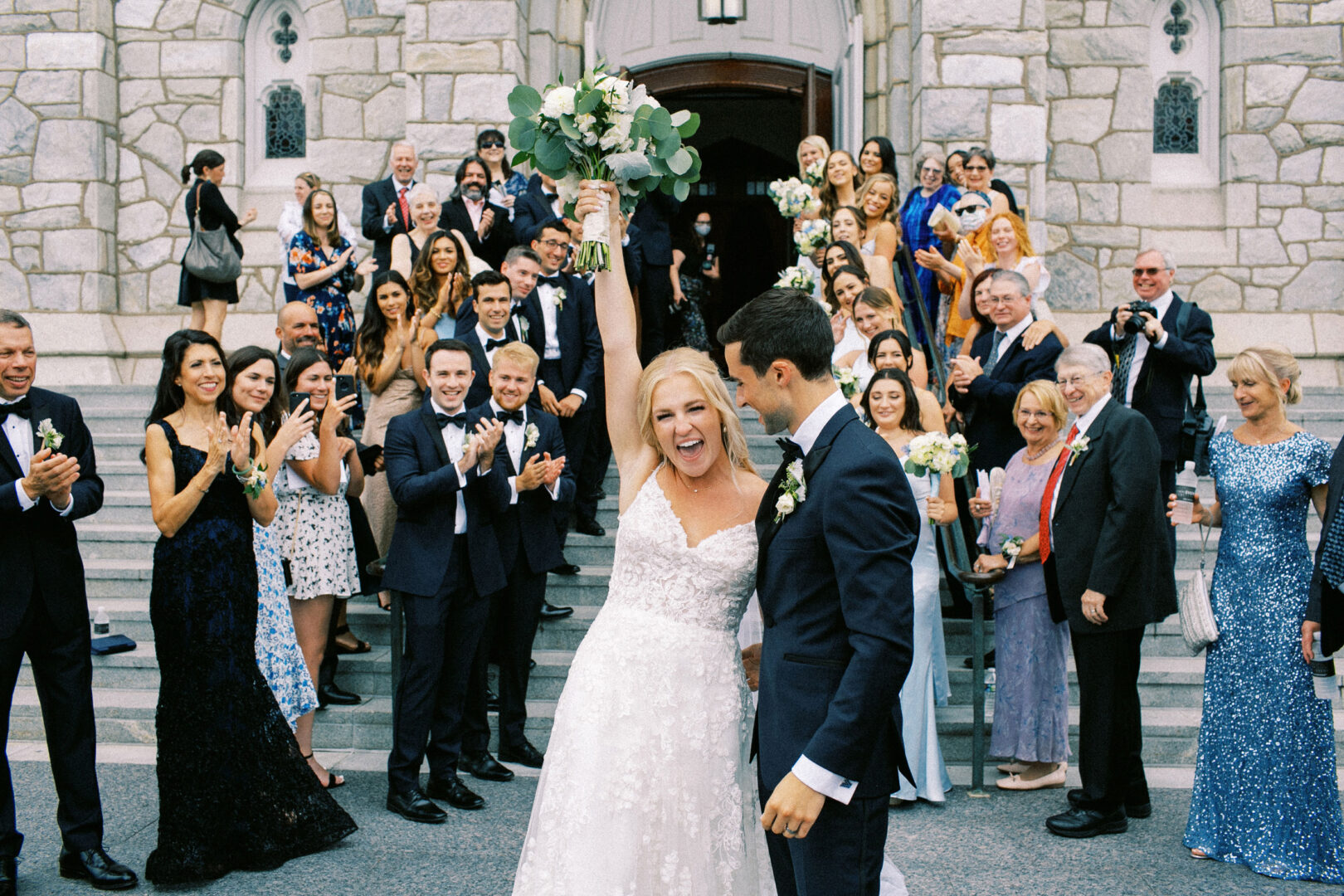 At a beautiful Normandy Farm wedding, the bride and groom celebrate outside a charming stone building, surrounded by cheering guests. The bride triumphantly holds her bouquet aloft, with everyone dressed in elegant formal attire.