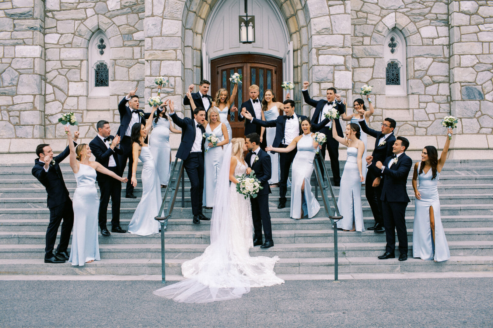 A bride and groom share a kiss on the steps, surrounded by a cheering wedding party in light blue and black attire, capturing the magic of their Normandy Farm Wedding against the backdrop of a stone building.