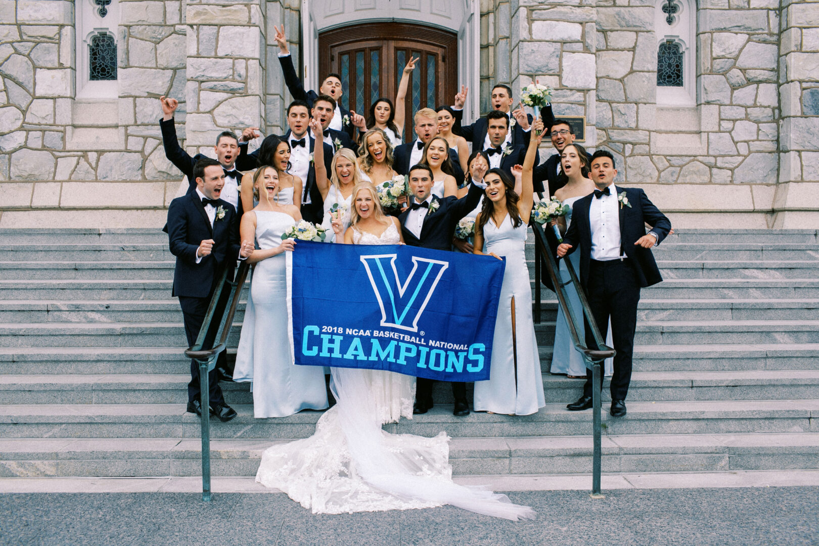 A group of people in formal attire stand on steps at Normandy Farm Wedding, proudly holding a banner reading "2018 NCAA Basketball National Champions.