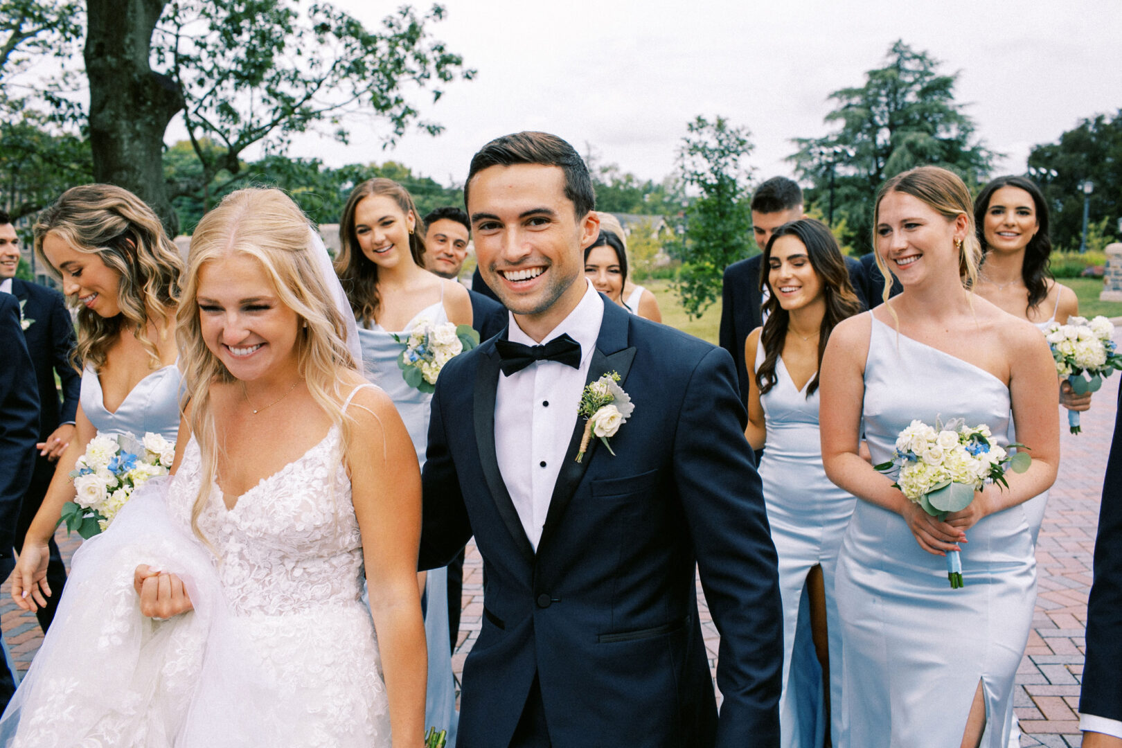 A bride and groom walk with their wedding party in matching outfits, holding bouquets. They appear joyful as they stroll the paved path at Normandy Farm Wedding venue, with lush trees providing a picturesque backdrop.