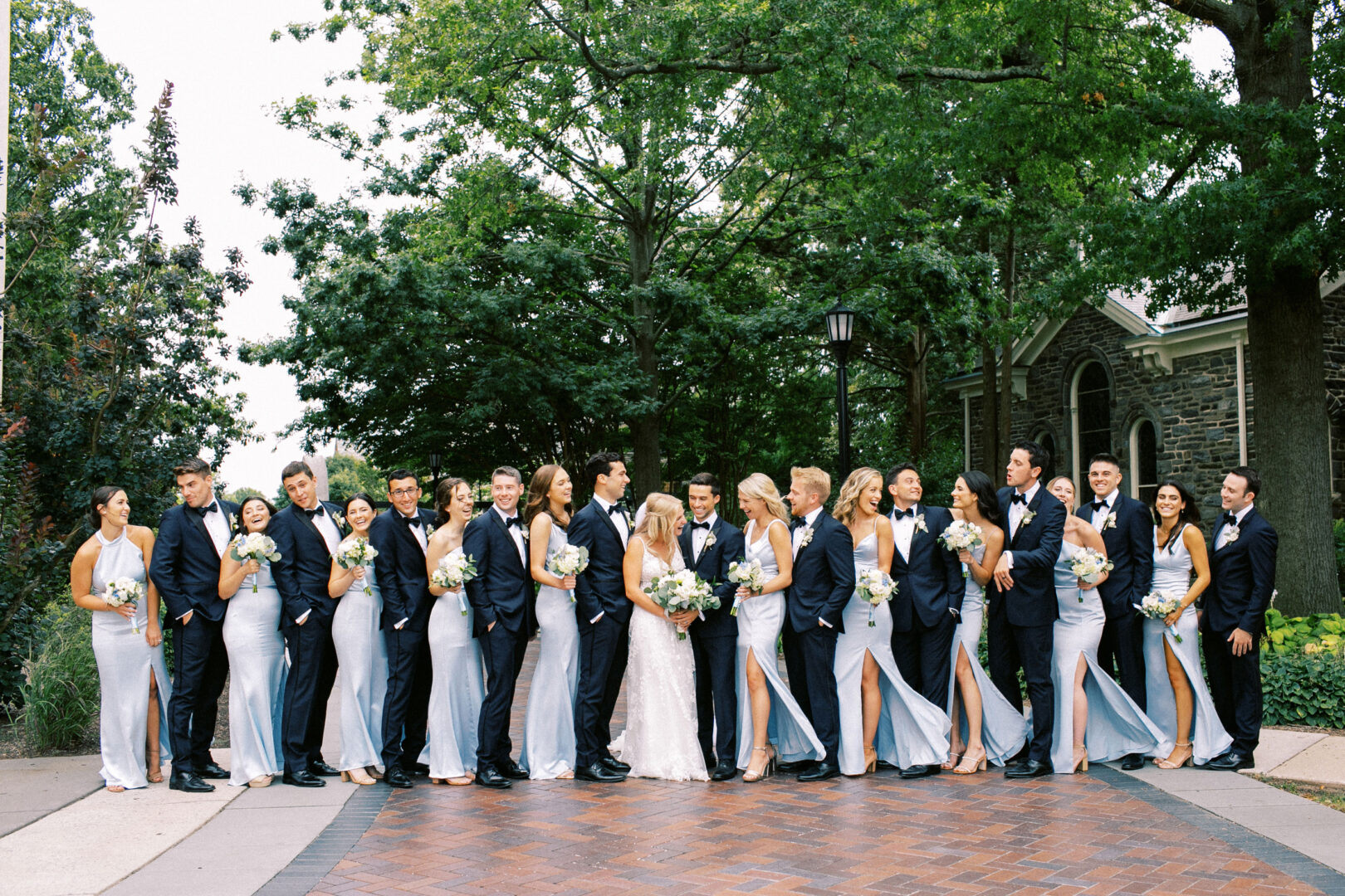 A large wedding party poses outdoors at Normandy Farm, with bridesmaids in light blue dresses and groomsmen in dark suits, next to trees and a stone building.