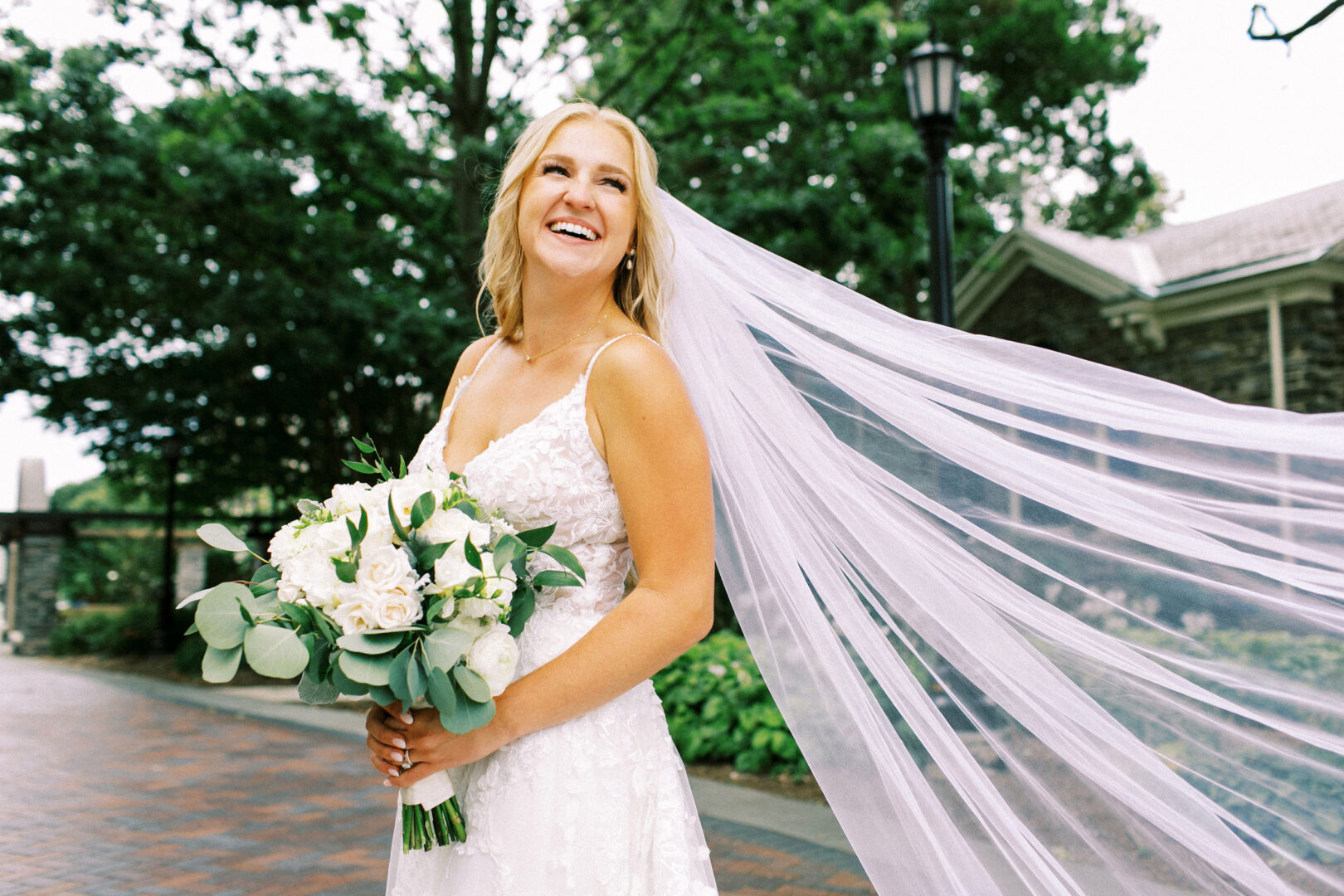 The bride, in her white wedding dress, smiles as her long veil flows in the wind at the picturesque Normandy Farm Wedding, surrounded by lush greenery and a charming building in the background.