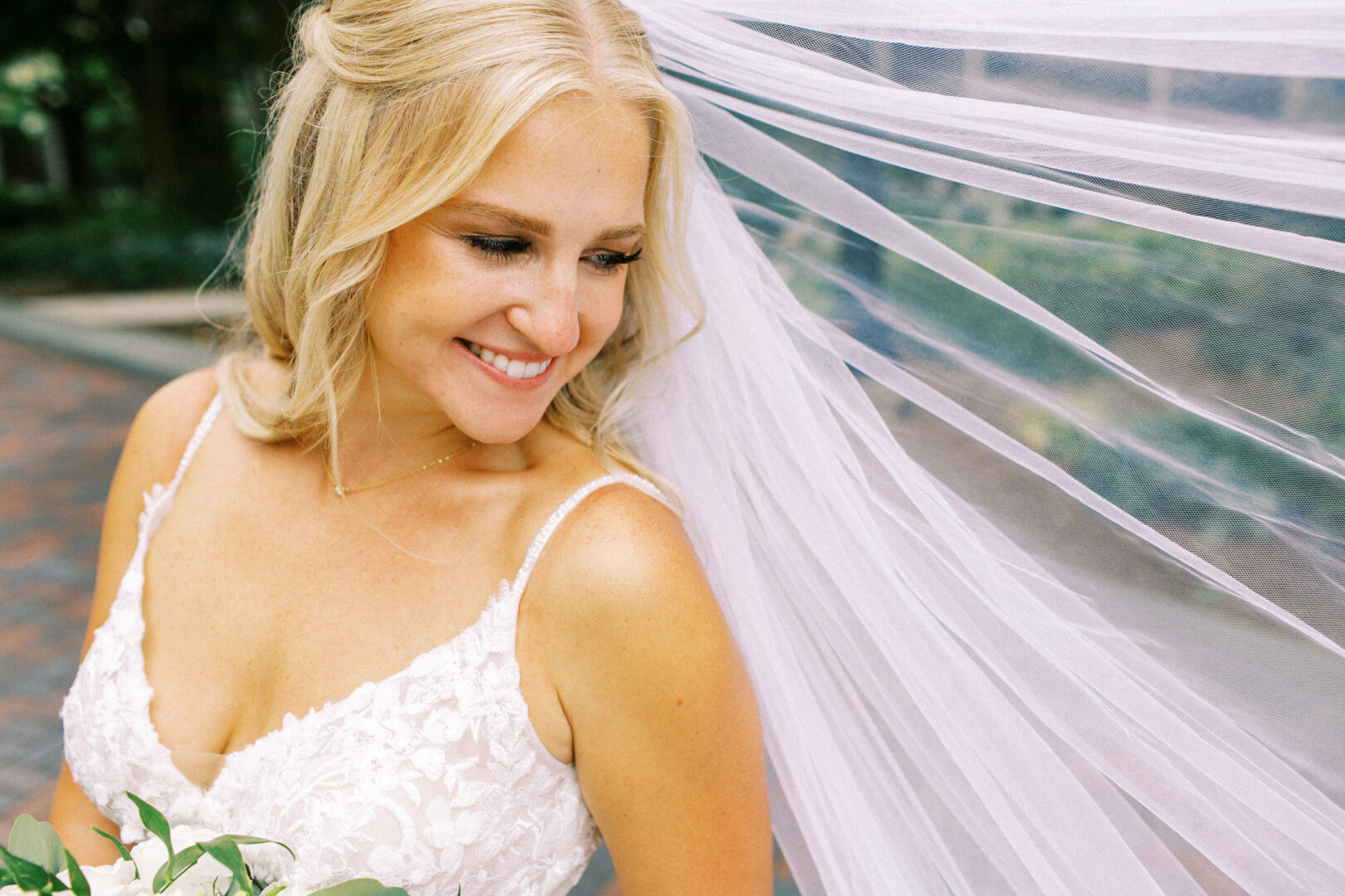 At a picturesque Normandy Farm wedding, the bride in a white lace dress and veil smiles radiantly while holding a bouquet outdoors.