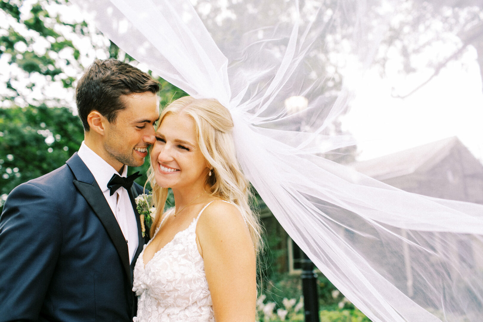 A bride and groom smile together outdoors at their enchanting Normandy Farm wedding, with the bride's veil flowing gracefully behind them.