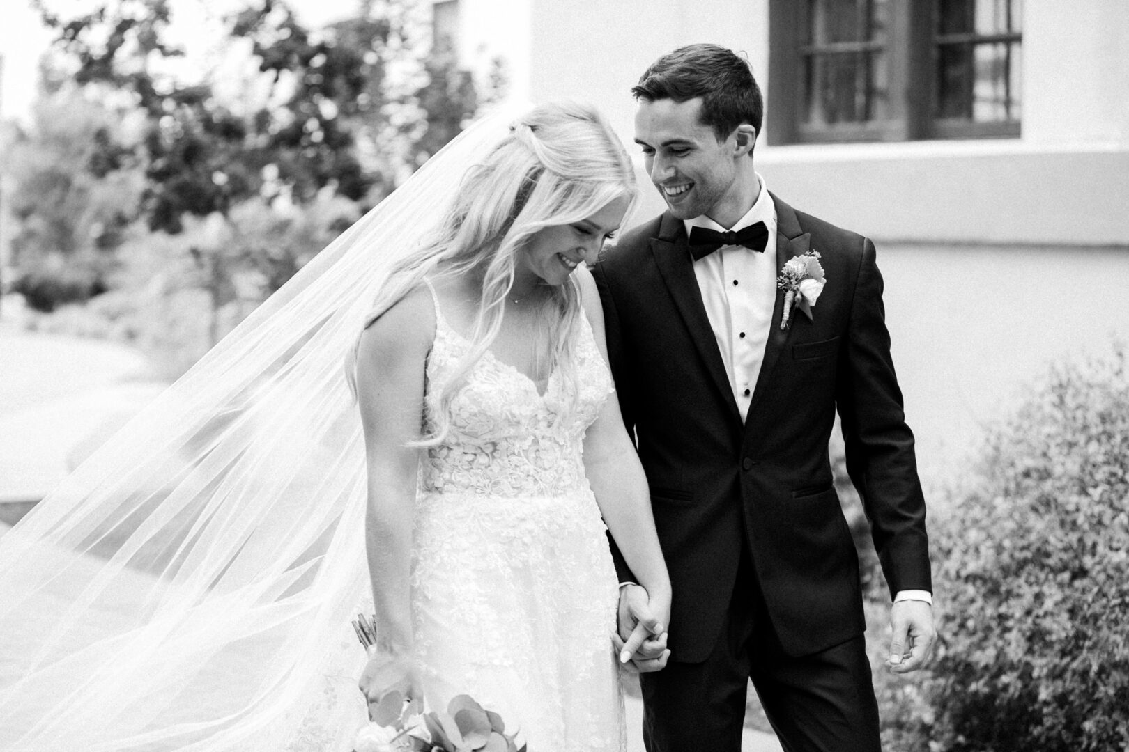 At a charming Normandy Farm wedding, a bride and groom walk hand in hand outdoors. The bride dazzles in a lace gown with a long veil, while the groom stands elegantly in his black tuxedo. Their smiles reflect the joy of this special day.