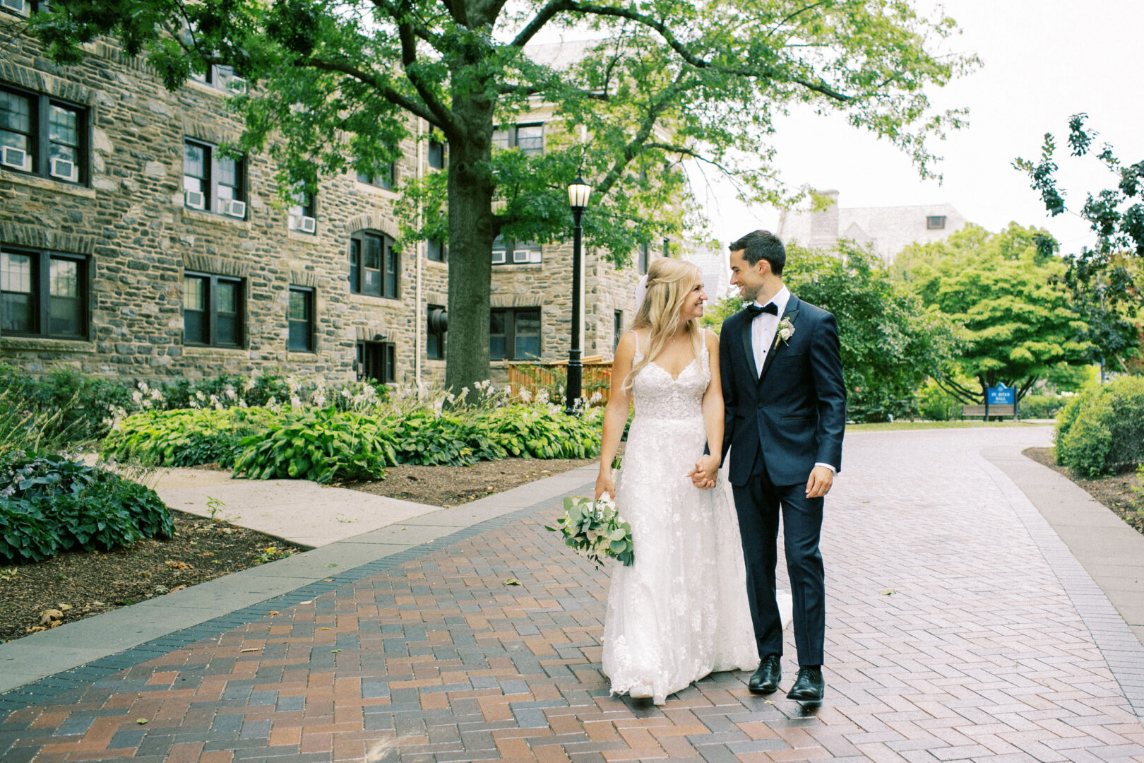 A bride and groom walk hand in hand on a brick path, surrounded by greenery and stone buildings, capturing the timeless charm of a Normandy Farm wedding.