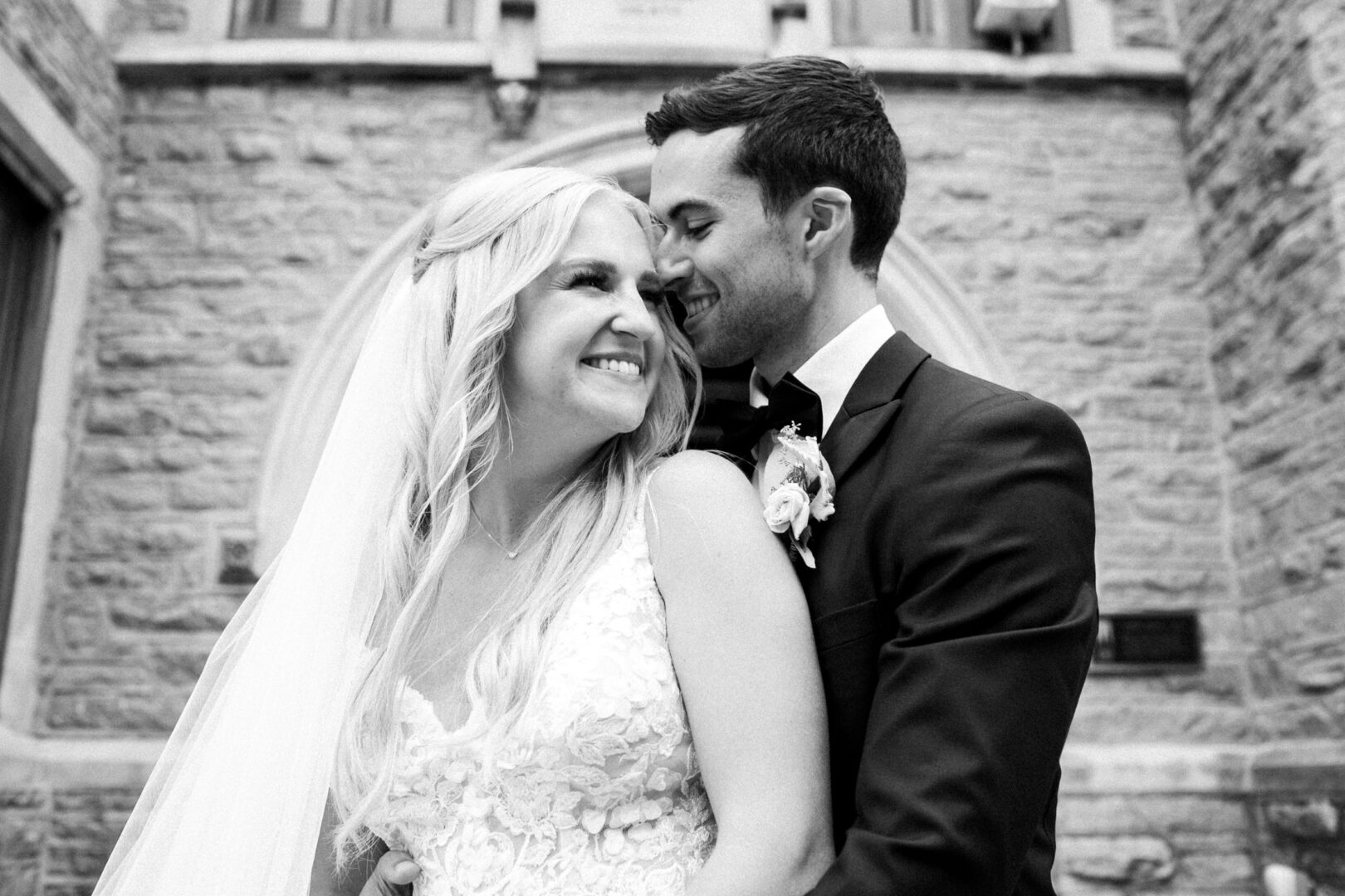 A smiling couple in wedding attire embrace in front of a stone building, capturing the essence of a Normandy Farm Wedding.