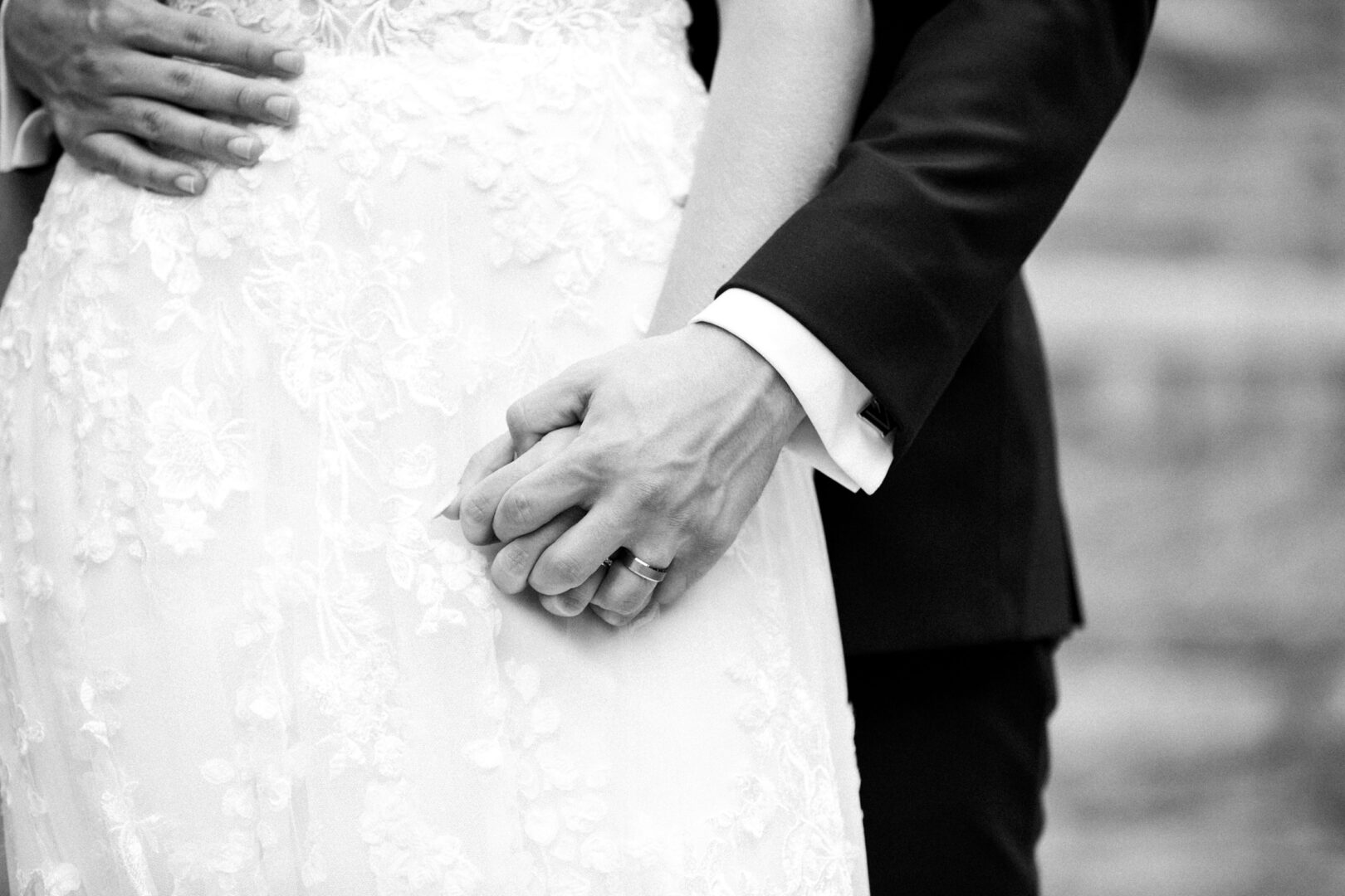 A couple in wedding attire holds hands at their Normandy Farm wedding, with the bride wearing a white lace dress and the groom in a dark suit.