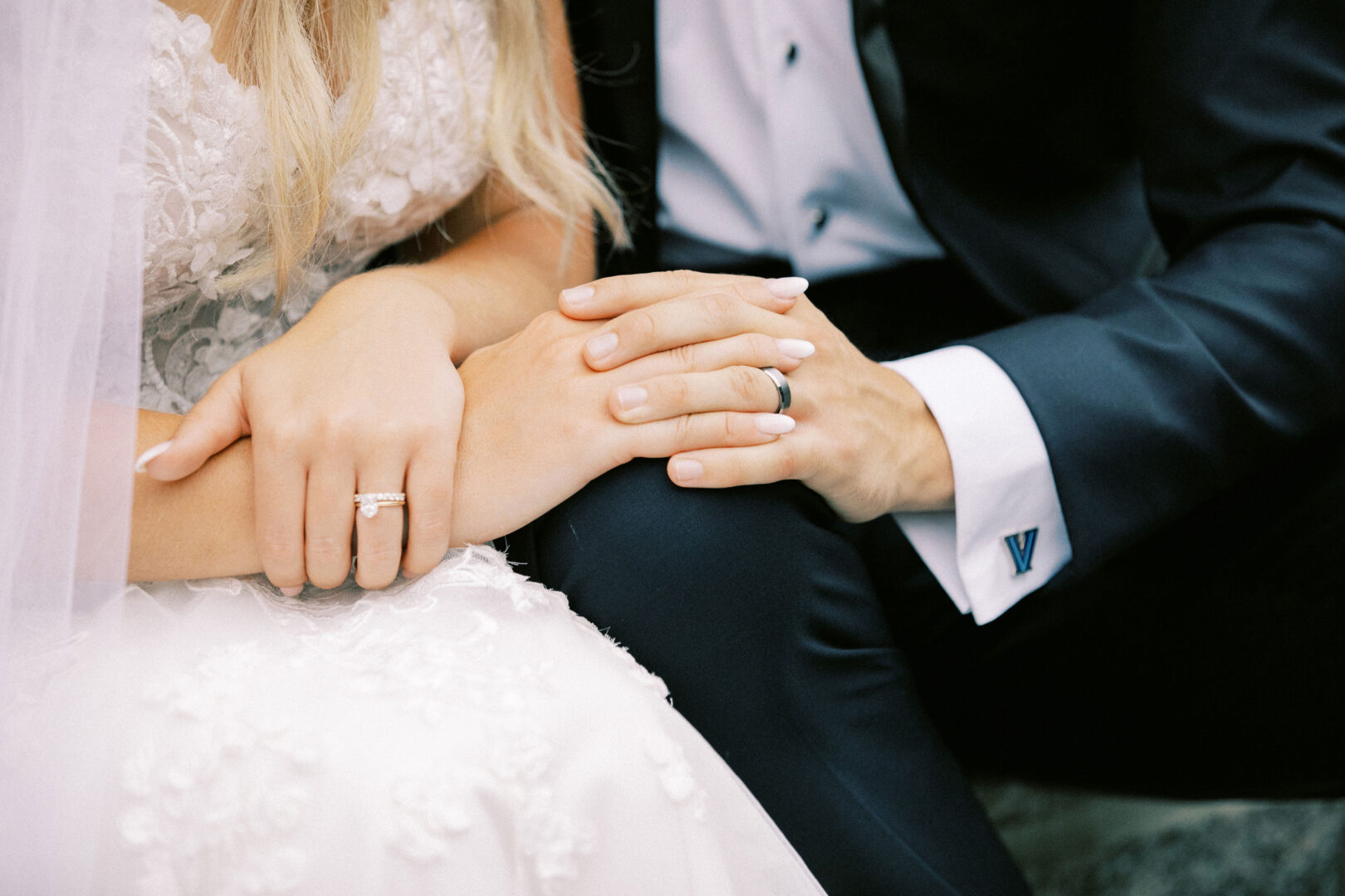 Close-up of a bride and groom seated at their Normandy Farm wedding, holding hands. The bride wears a white lace dress and veil with a ring on her finger; the groom is in a dark suit with a white shirt.