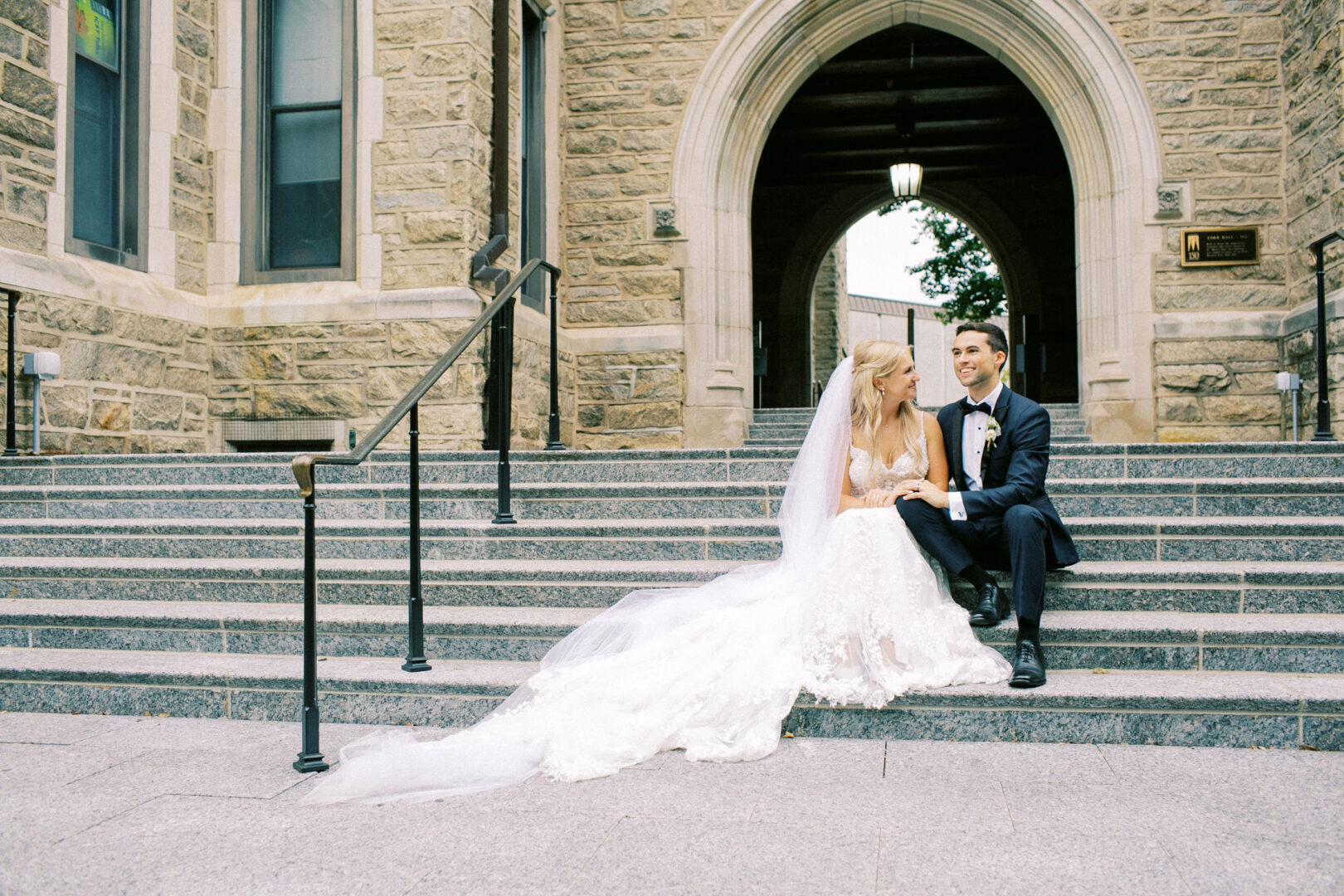 A bride and groom sit on stone steps at Normandy Farm's arched entrance, the bride radiant in a white gown and the groom sharp in a black tuxedo.