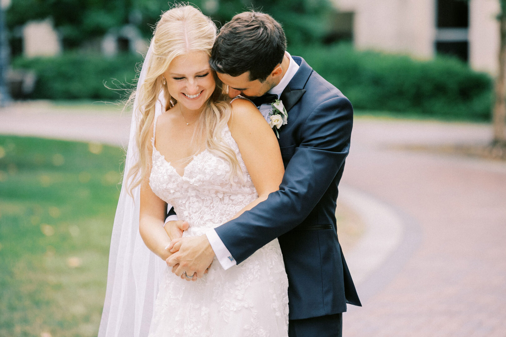 The couple, celebrating their Normandy Farm Wedding, embraces outdoors on a picturesque pathway. The bride dazzles in her white lace gown and veil, while the groom looks dapper in a dark suit with a boutonniere. They stand smiling under the sun’s warm embrace.