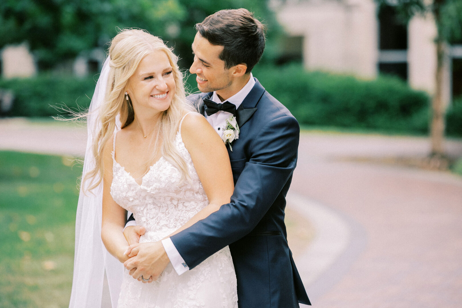 The bride and groom embrace and smile outdoors on a sunny day at their Normandy Farm wedding, surrounded by lush greenery and winding paths.