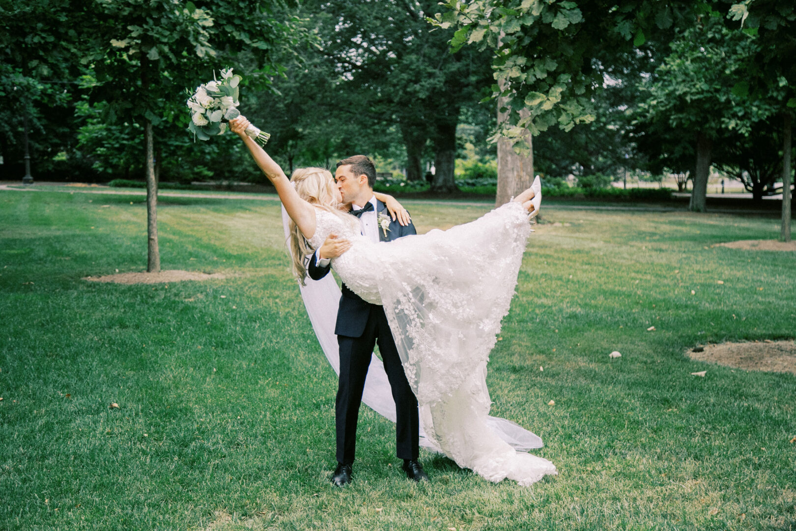 At their Normandy Farm wedding, a groom in a tuxedo lifts his bride in a lace gown as they pose under trees in the grassy park. The bride joyfully holds her bouquet aloft.