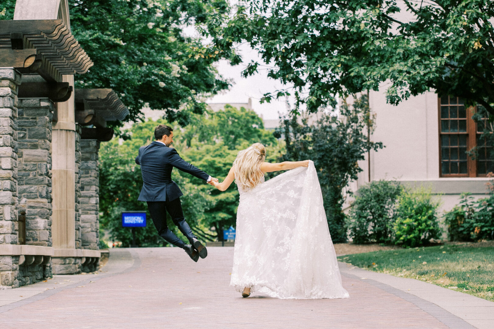 The bride and groom joyfully hold hands and leap along a tree-lined path, capturing the enchanting spirit of their Normandy Farm wedding in a picturesque park setting.