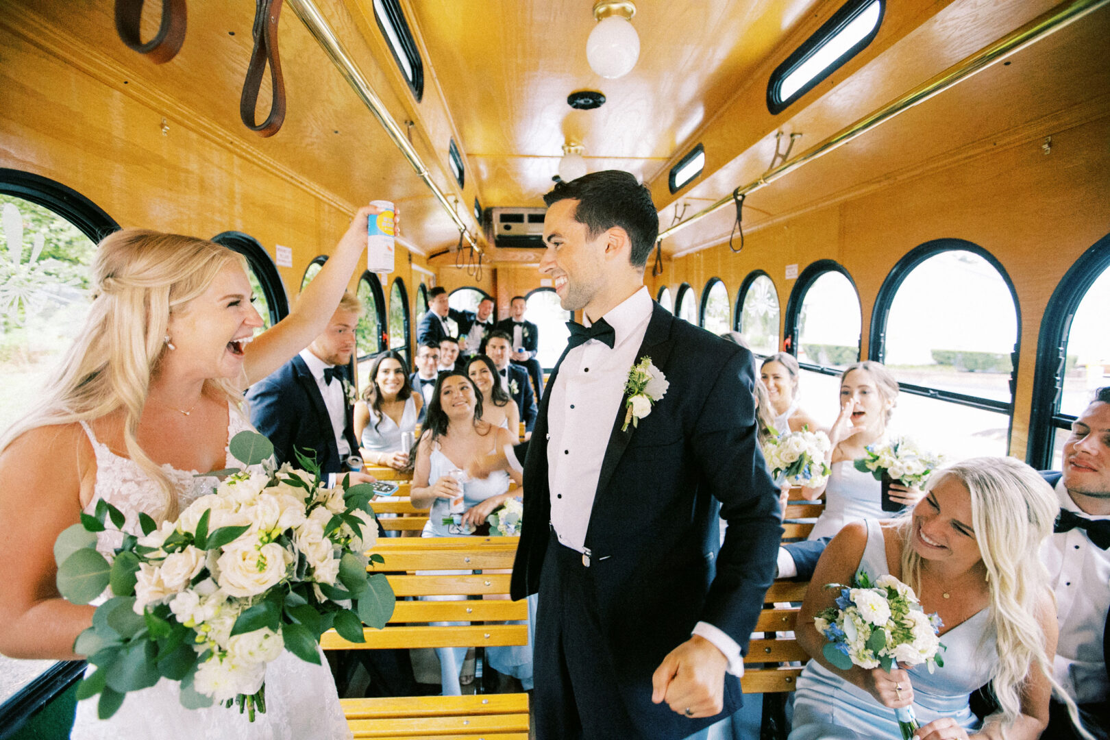 At a charming Normandy Farm Wedding, the bridal party dressed in elegant formal attire joyfully rides a trolley. The bride exuberantly holds up a can amid laughter and smiles from delighted guests.