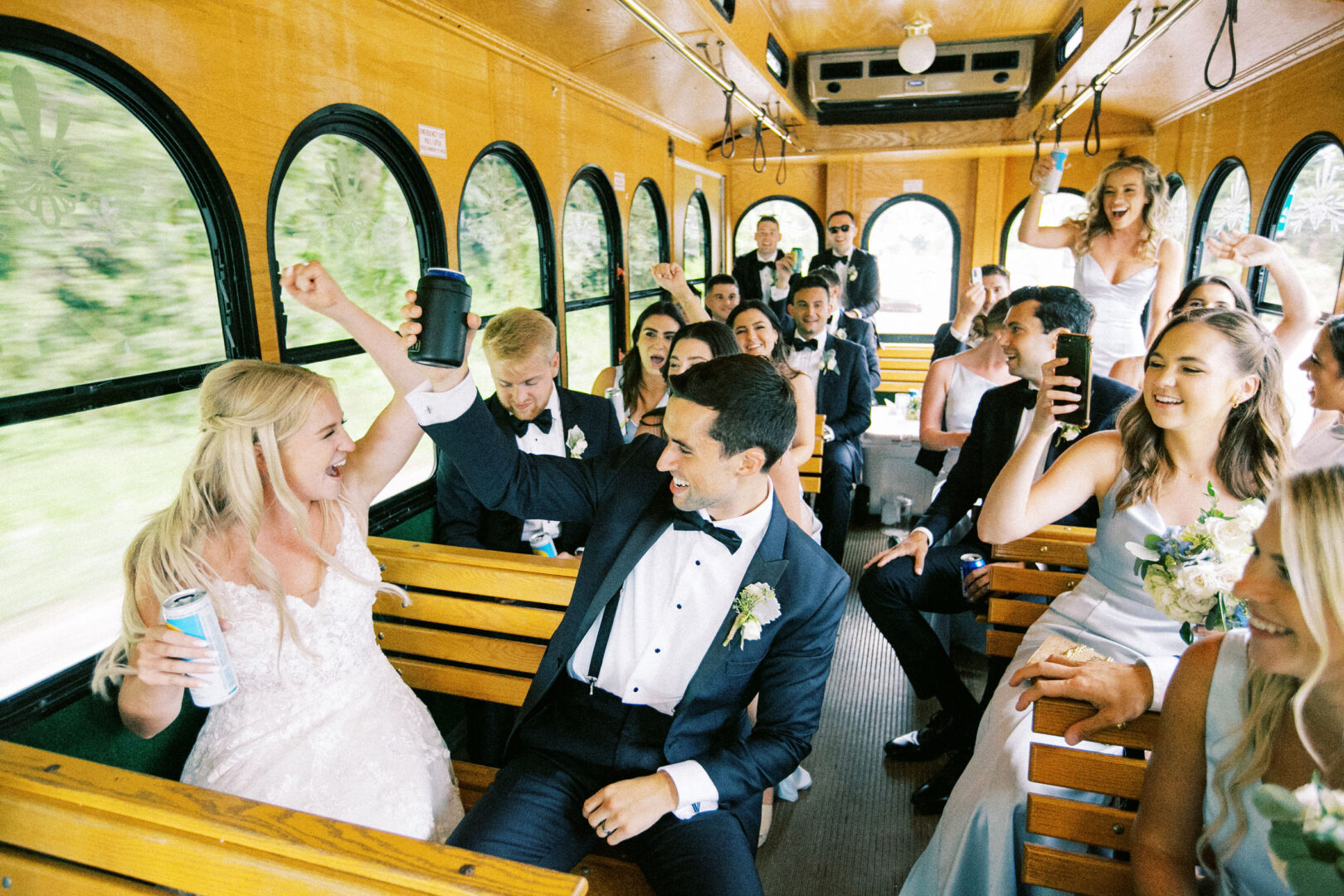 A group of people in formal attire celebrates on a party bus, raising cans and smiling, as they joyously reflect on the beautiful Normandy Farm wedding.