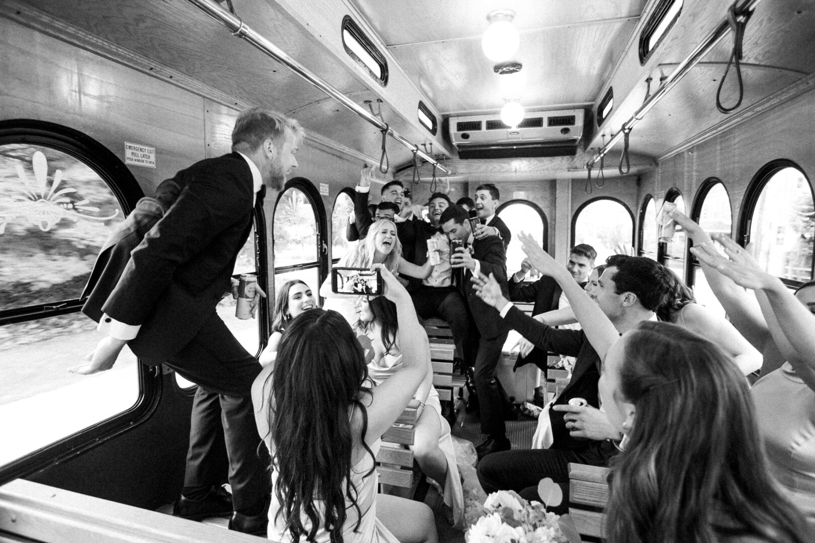 A group of people in formal attire celebrate energetically inside a trolley bus, capturing the moment with photos and raising their hands, as if reliving a Normandy Farm Wedding.